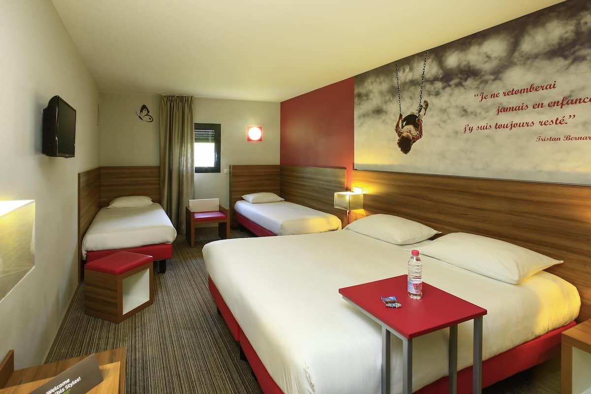 Une chambre d’hôtel avec deux lits, une petite table sur laquelle se trouve une bouteille, une télévision et un mur orné d’une citation.