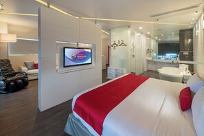 A modern hotel room with a large bed, a TV mounted on the wall, a seating area with a massage chair, and a separate bathroom with a mirror and sink. - Ramada Encore by Wyndham Puebla (Coronango)