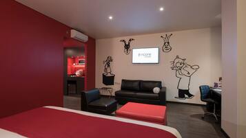 Una habitación de hotel moderna con una pared de acento roja, una cama grande, un sofá de cuero negro, un escritorio pequeño y una pantalla de televisión que muestra el logotipo de Encore Hotels.