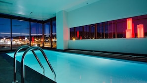 A modern indoor pool with clear glass walls, a metal ladder, and a view of a cityscape at night.