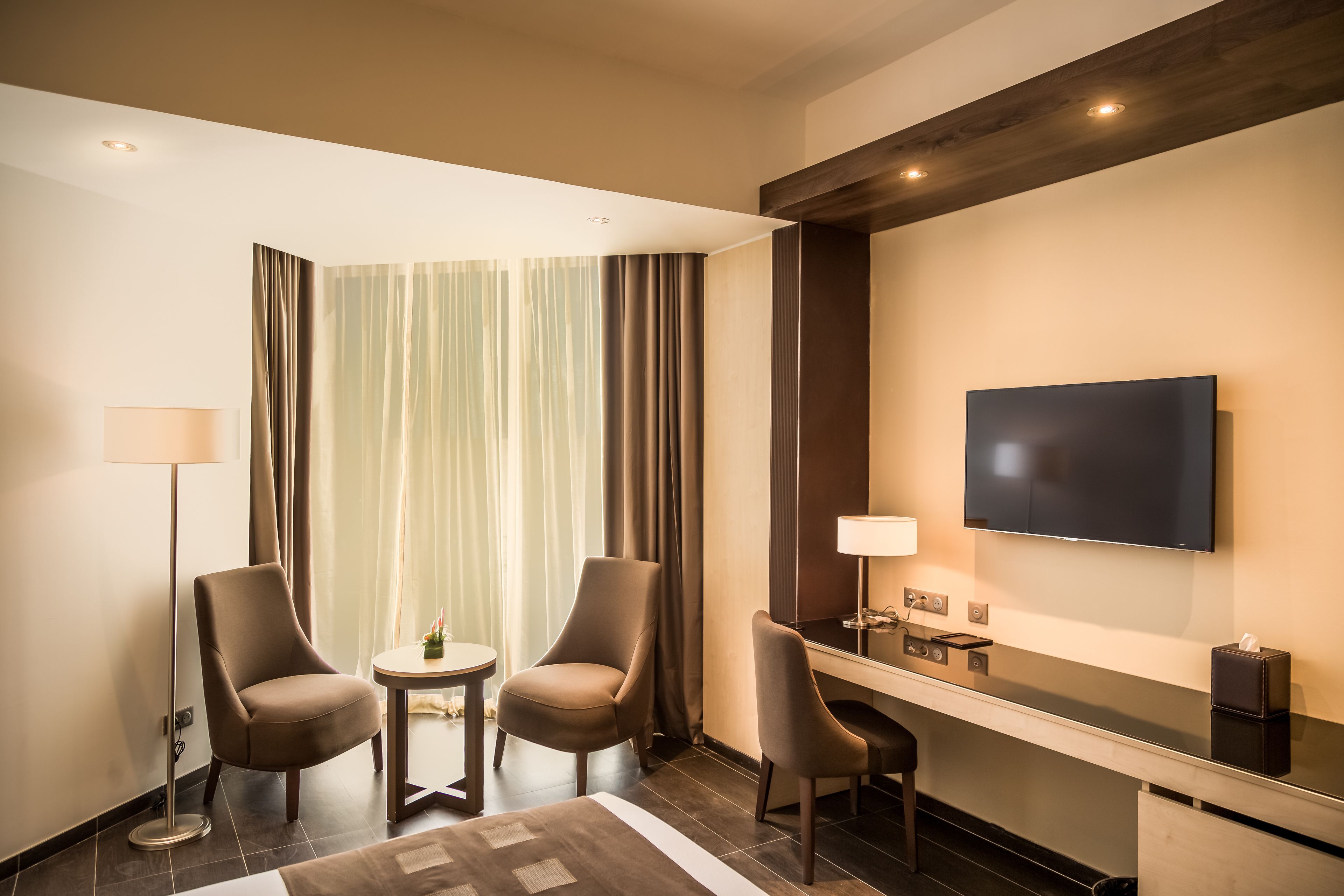 A hotel room with two armchairs, a small table, a flat-screen TV, a desk with a lamp, and a large window with curtains.