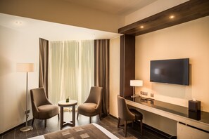 A hotel room with two armchairs, a small table, a flat-screen TV, a desk with a lamp, and a large window with curtains. - Azalai Hôtel Abidjan (Abidjan)