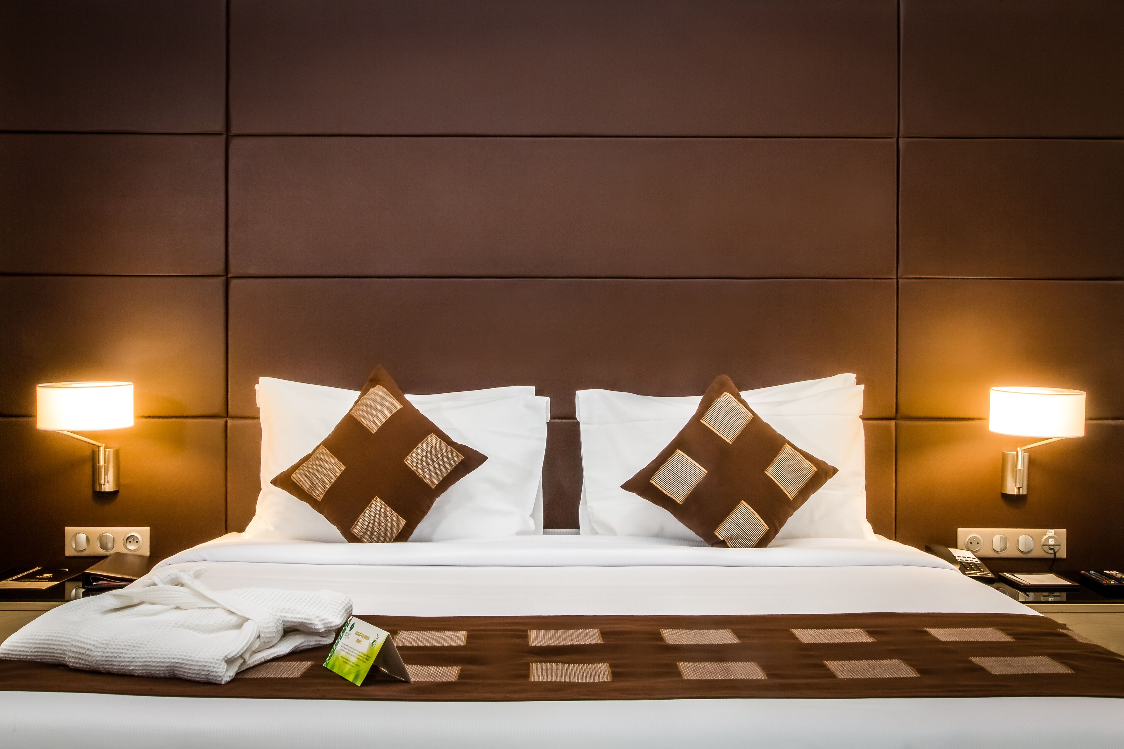 Una cama bien tendida con sábanas blancas y almohadas marrones, dos lámparas de noche, un control remoto y un par de toallas blancas sobre la cama.