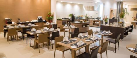 A modern hotel dining area with multiple tables and chairs, a central island with a coffee machine, and a buffet setup.