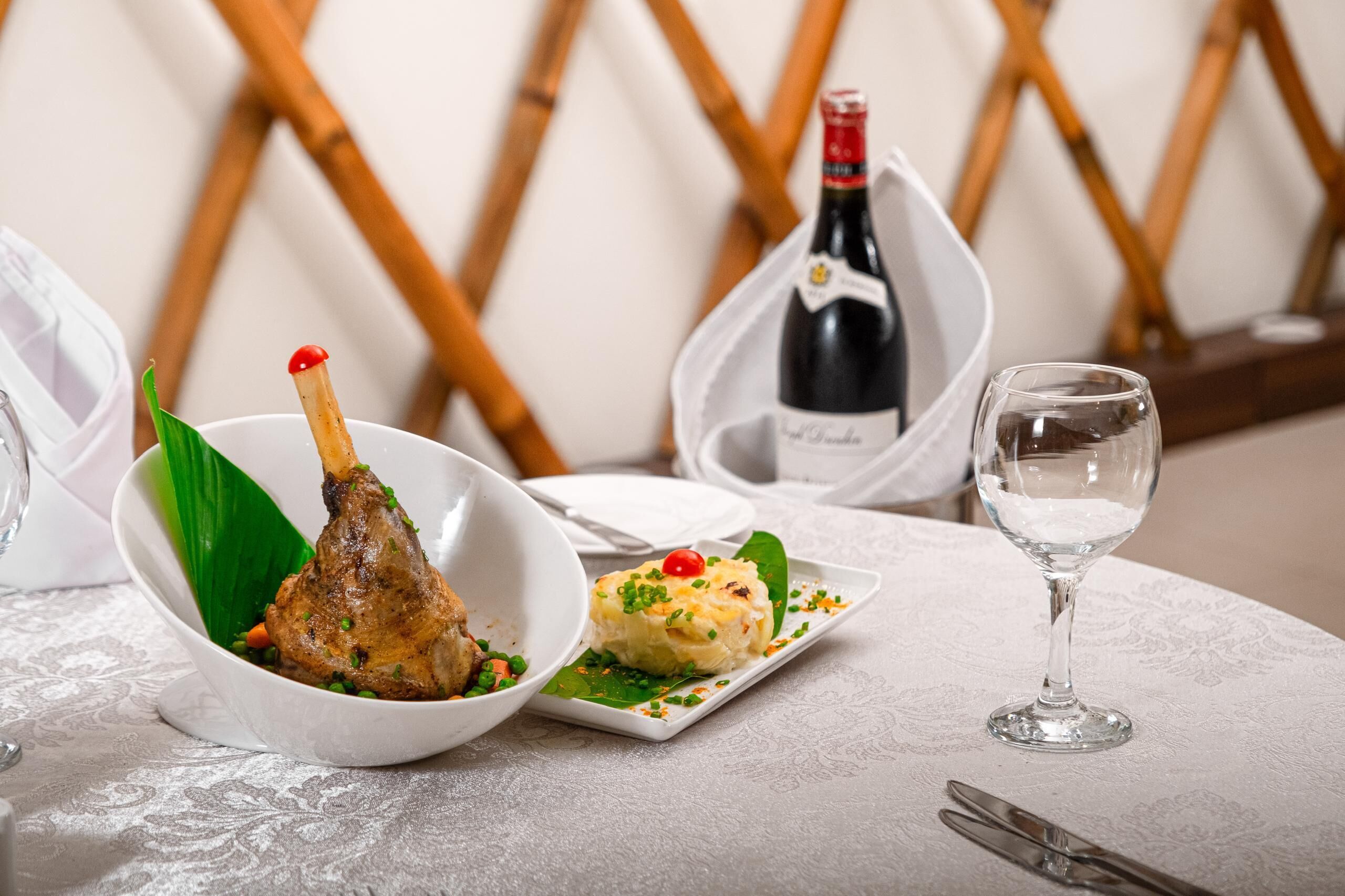 A table setting with a lamb shank, a small dish of mashed potatoes, a wine bottle, and a wine glass.