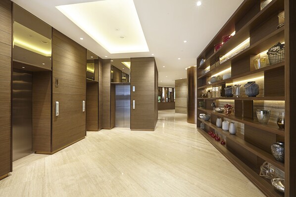 A modern hotel corridor with wooden paneling, a central elevator, and a display of decorative vases on shelves.
