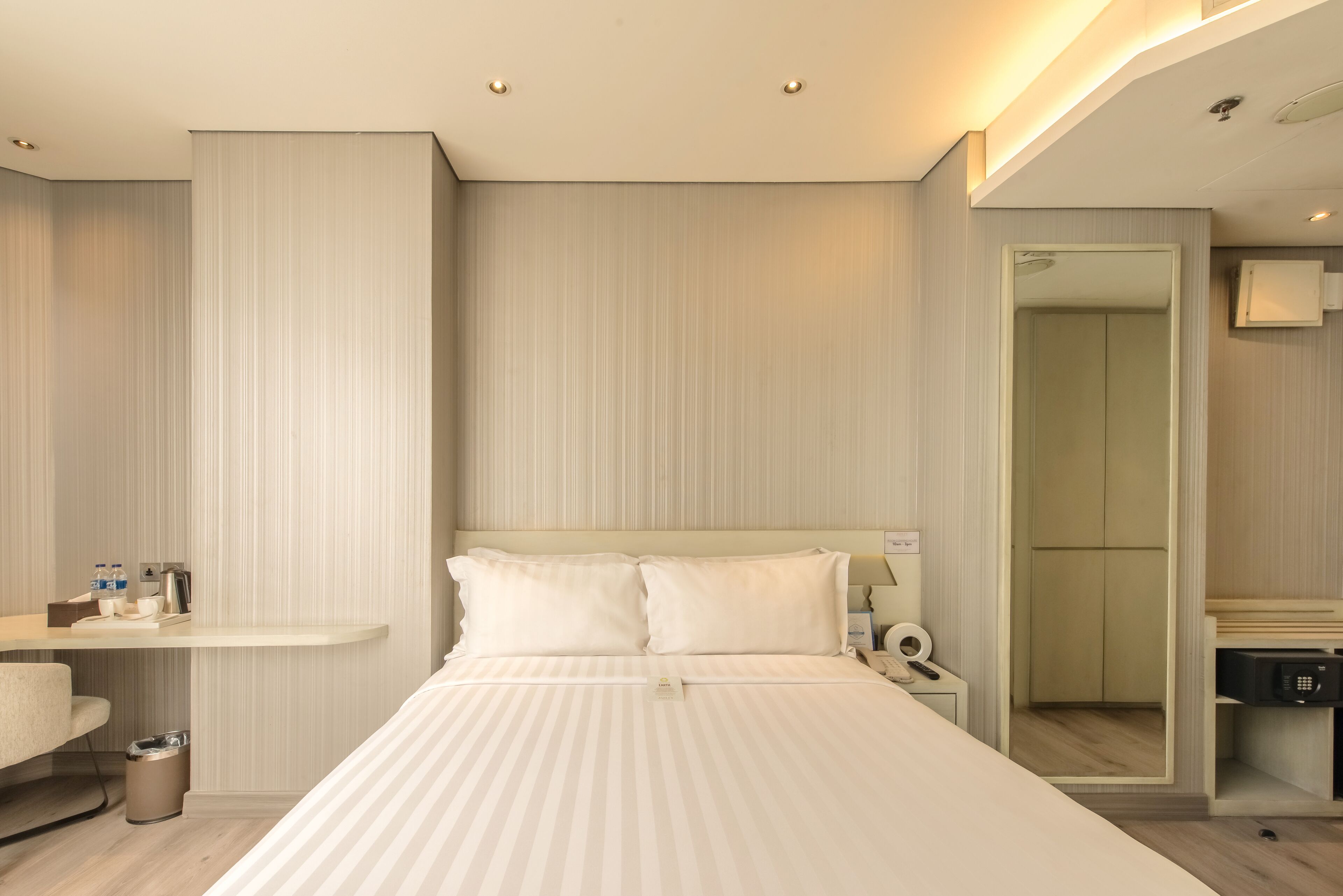 A modern hotel room with a large bed, a bedside table with a lamp, a small trash bin, and a wardrobe with a mirror.