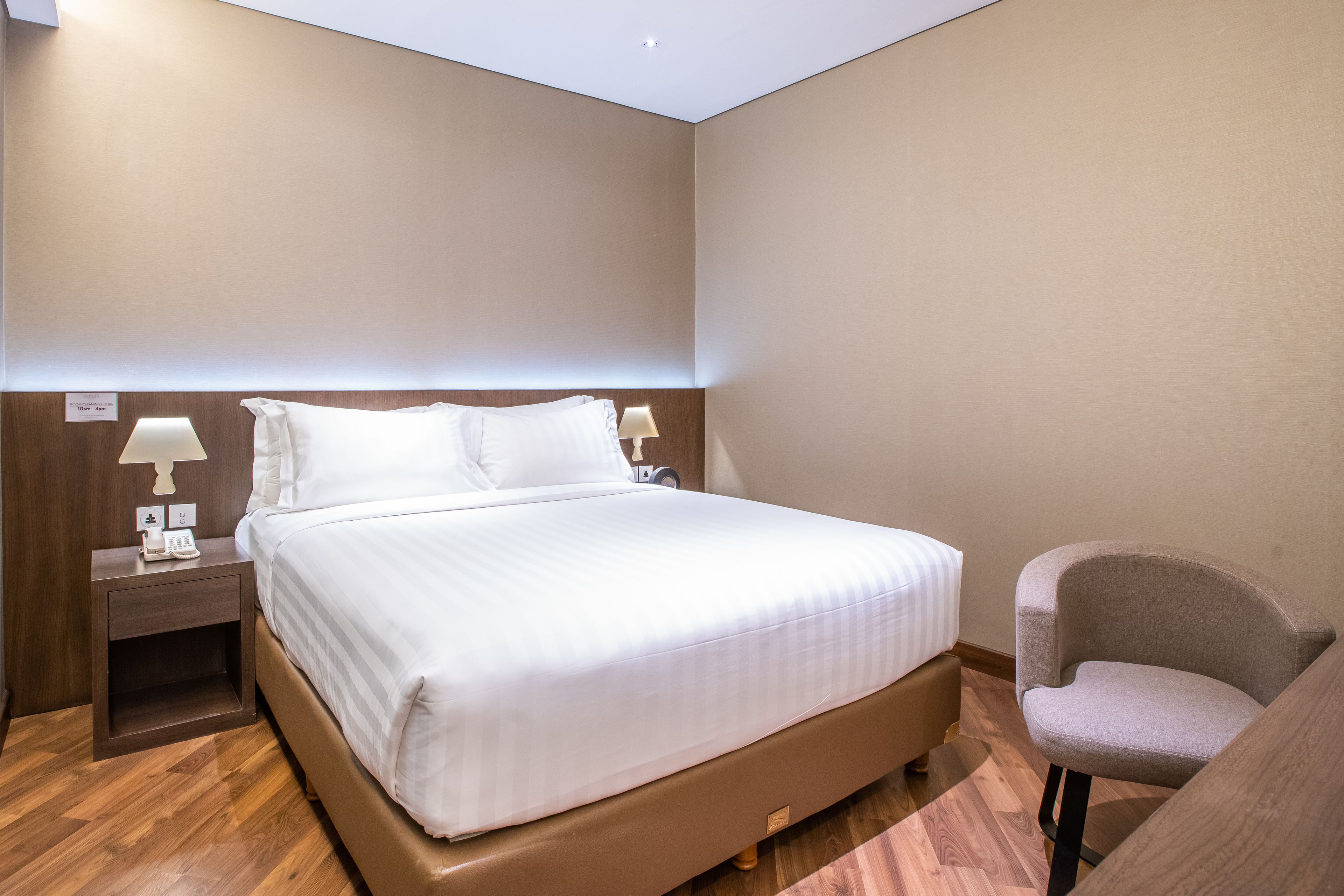 A neatly made bed with white linens, a bedside table with a lamp, and a chair in a modern hotel room.
