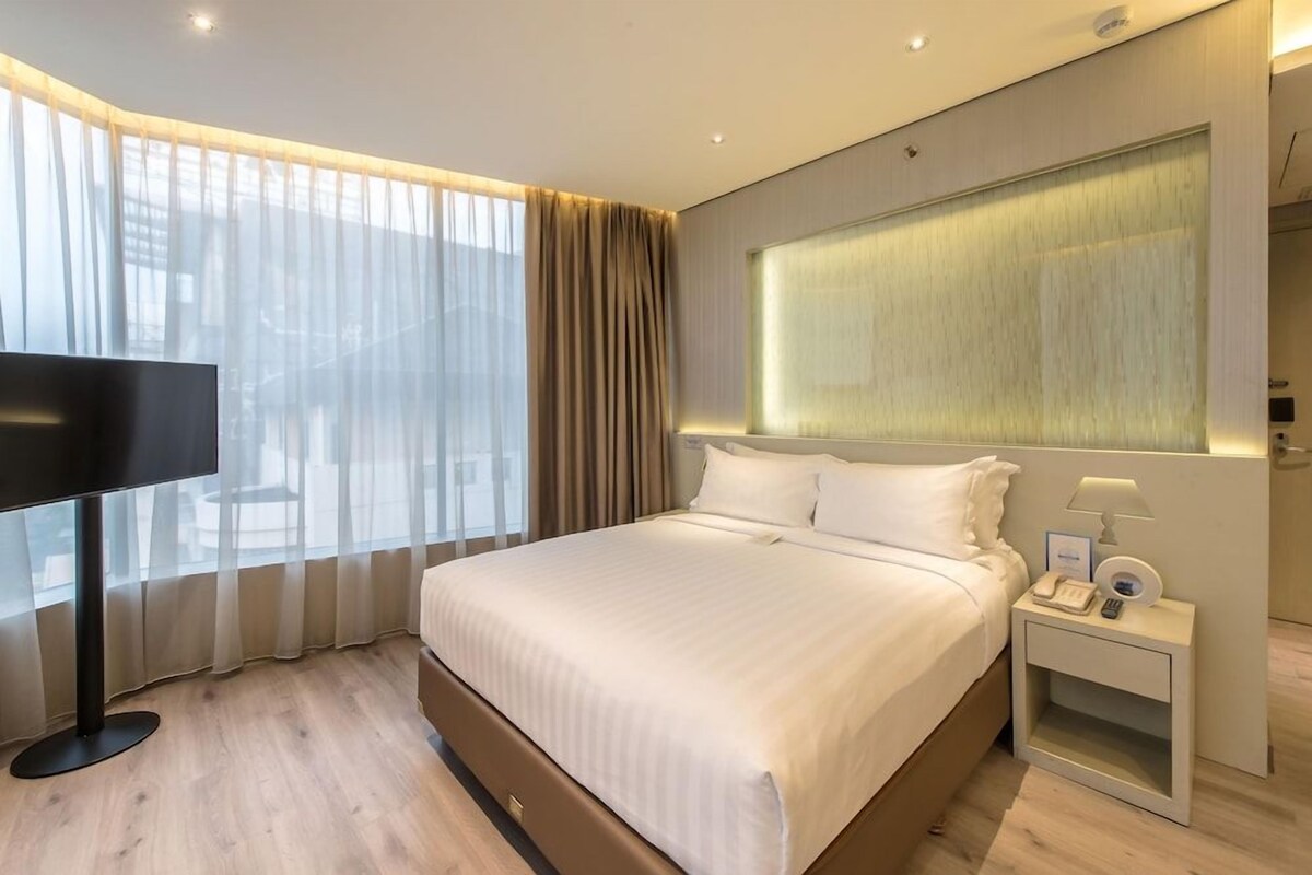A modern hotel room with a large bed, a TV, a bedside table with a lamp, and a window with sheer curtains.
