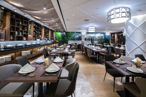 A restaurant with multiple dining tables set with white tablecloths and candles, surrounded by shelves with various items and a green mural on the wall. - Ashley Wahid Hasyim Jakarta (Jakarta)