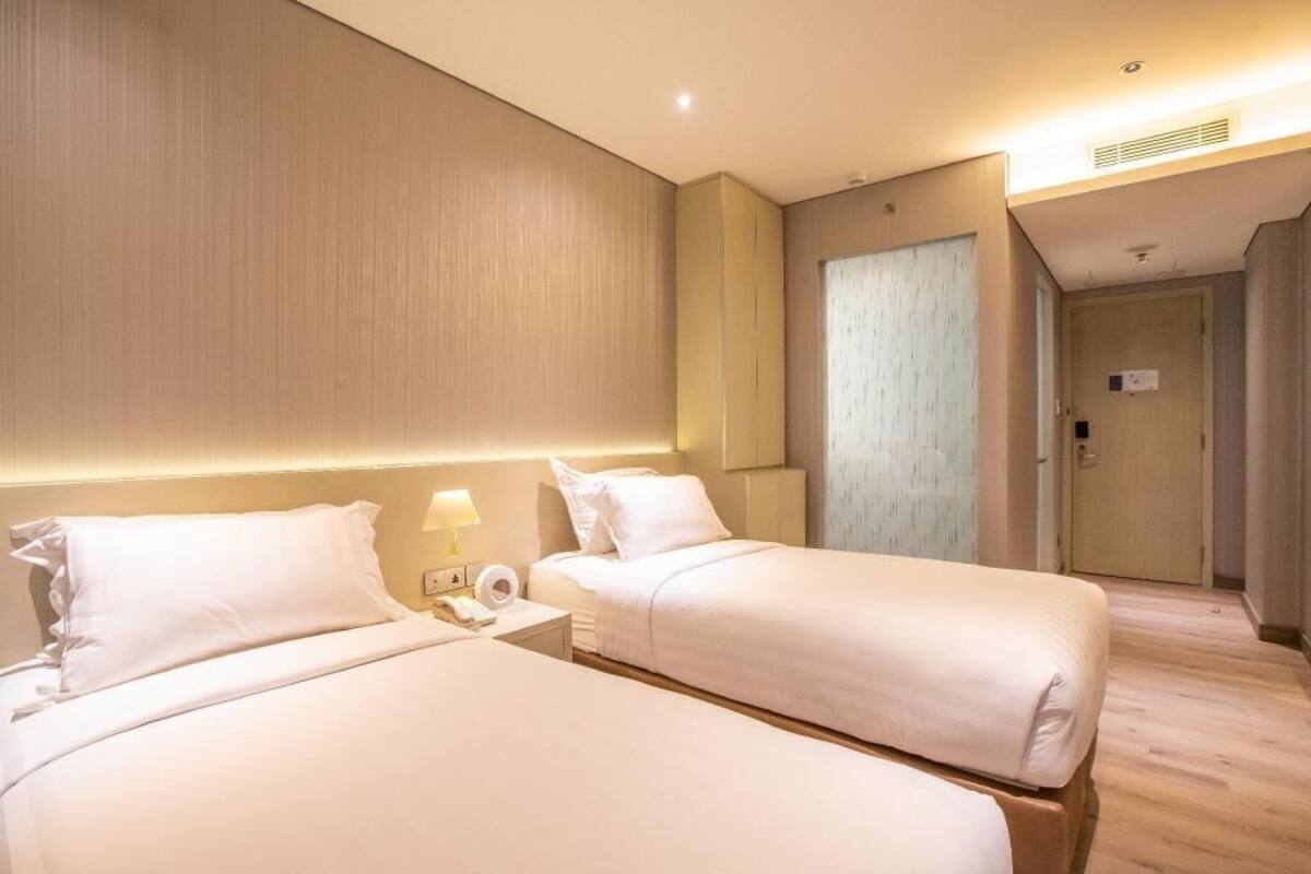 A hotel room with two beds, a bedside table with a lamp, and a door leading to another room.