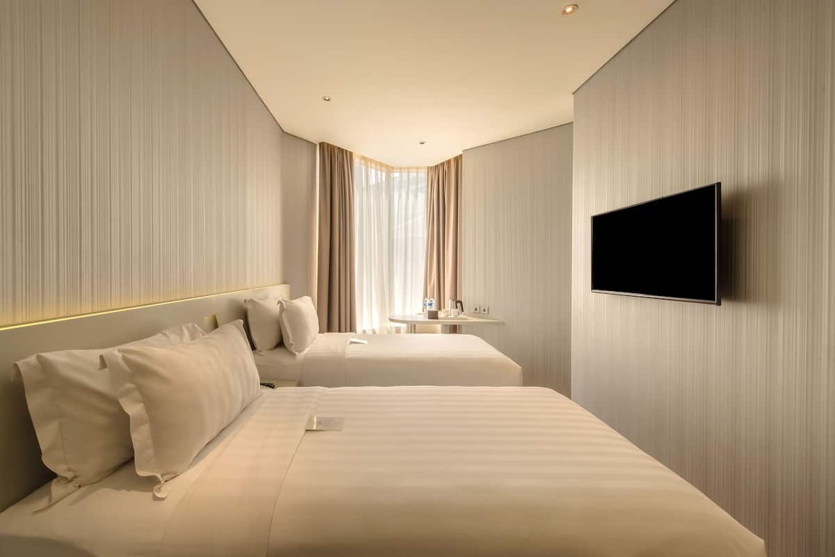 A hotel room with two beds, a TV mounted on the wall, a small table, and a window with curtains.