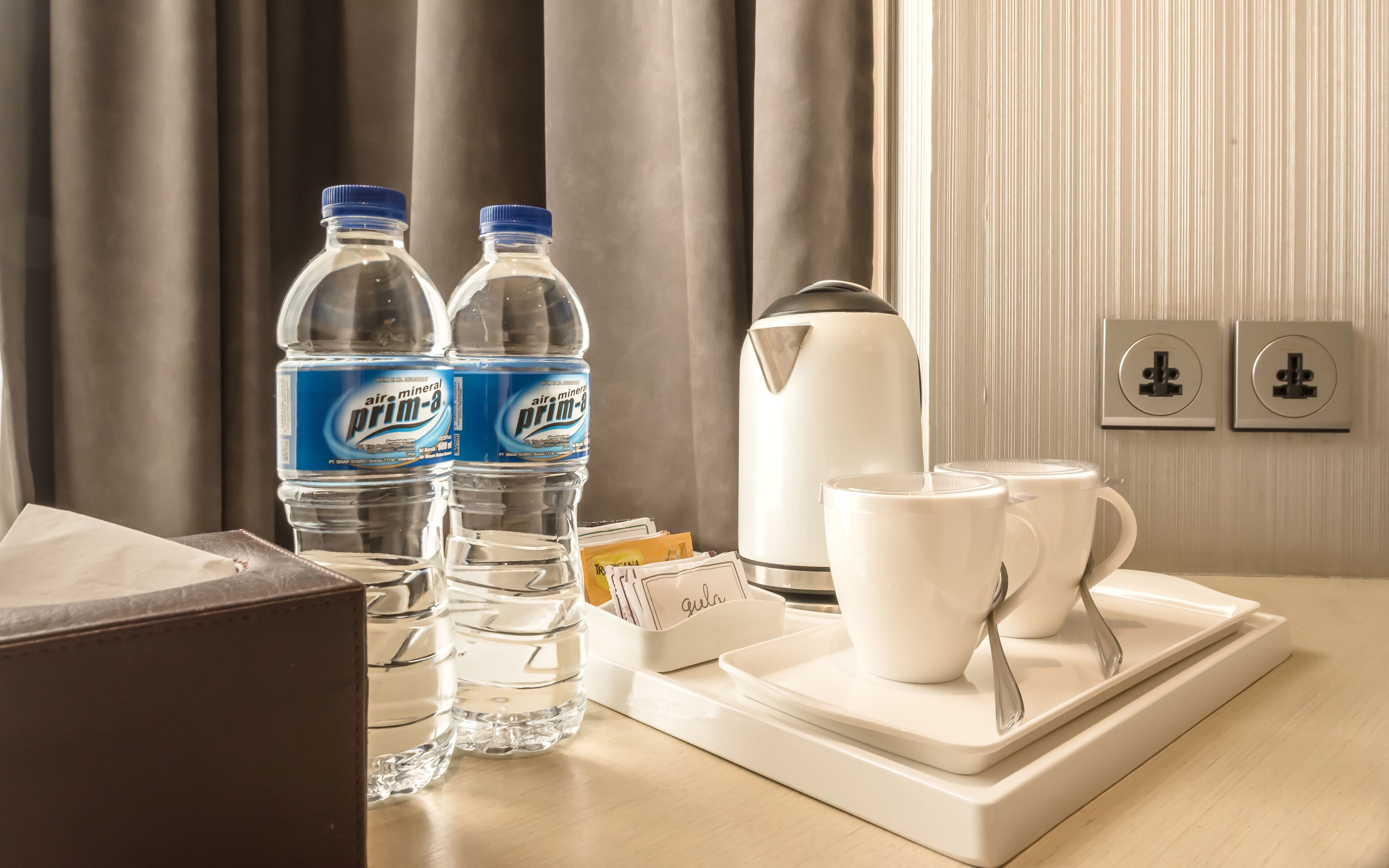 A hotel room with a table set with two bottles of air mineral prim-a water, a white electric kettle, two white mugs, and a tray with a spoon.