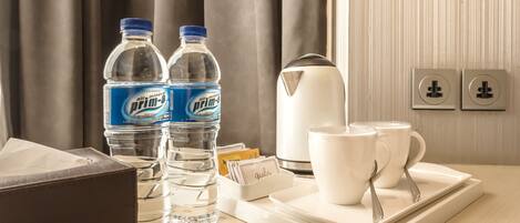 A hotel room with a table set with two bottles of air mineral prim-a water, a white electric kettle, two white mugs, and a tray with a spoon.