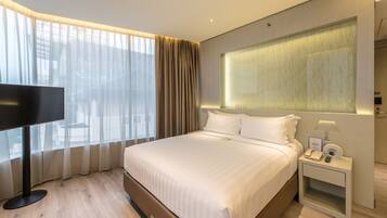 A modern hotel room with a large bed, a flat-screen TV, a bedside table with a lamp, and a large window with sheer curtains.