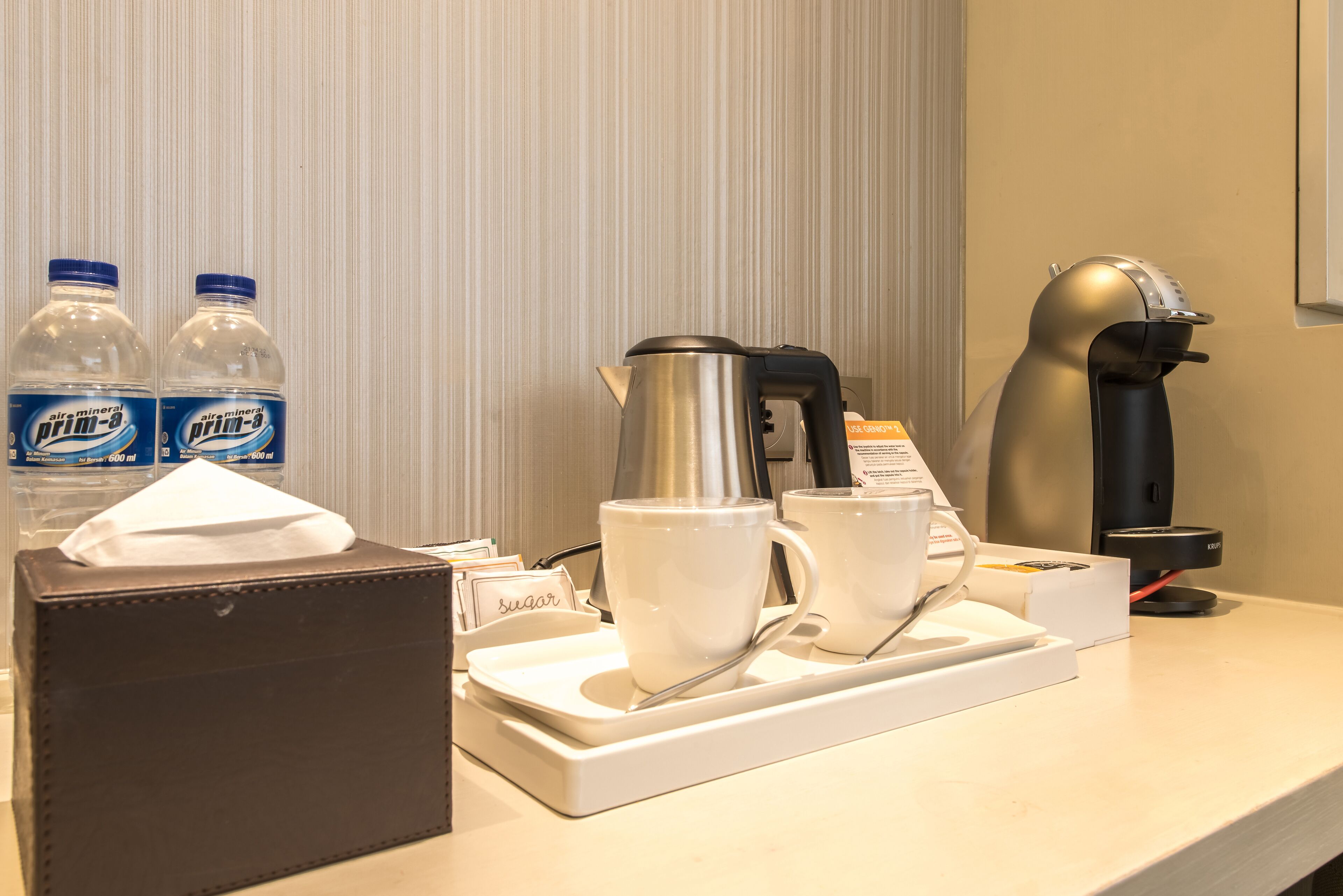Preparación de desayuno en una habitación de hotel, con una cafetera, dos tazas, una caja de pañuelos y dos botellas de agua.