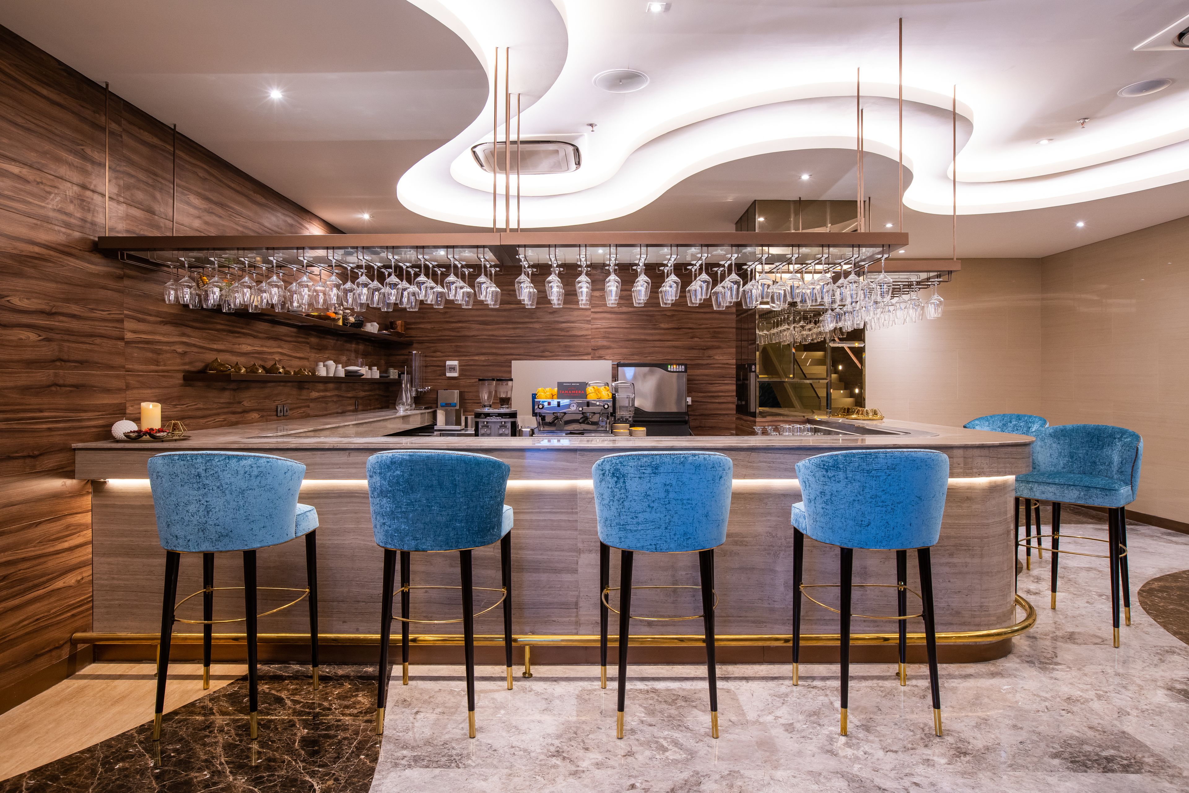 A modern bar area with a marble floor, blue bar stools, and a glassware display.