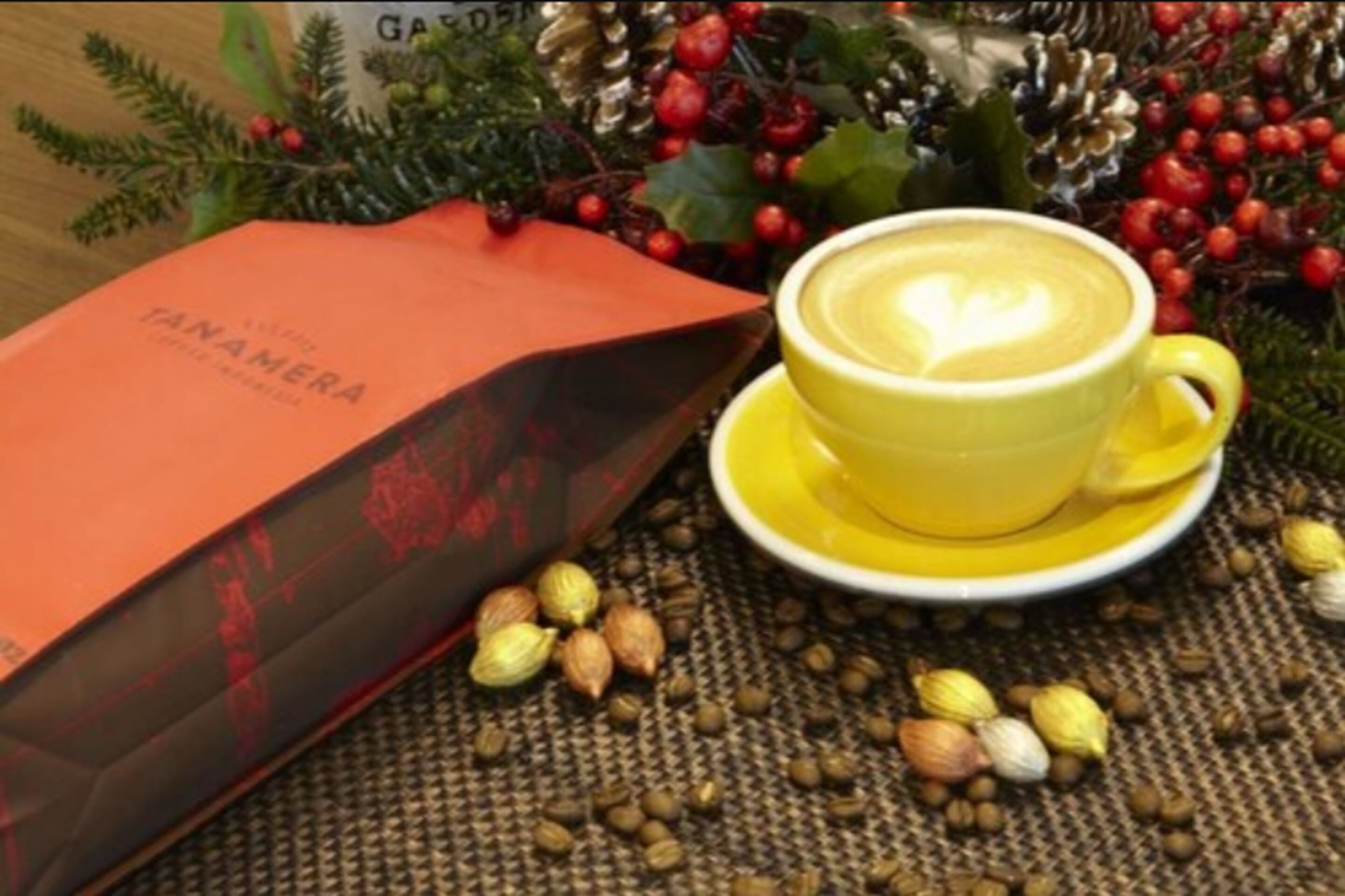 Una taza amarilla de café con un diseño de corazón en la espuma, sobre un platillo amarillo, rodeada de nueces esparcidas y una bolsa roja con la etiqueta "TANAMERA".
