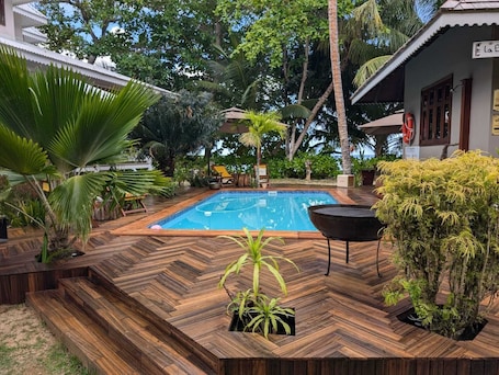 A pool area with wooden decking, surrounded by tropical plants and a palm tree.
