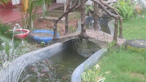 Un petit pont enjambant un bassin agrémenté de jeux d’eau, entouré de verdure et bordé d’un mur de pierres.
