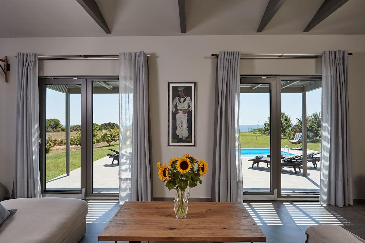 A room with a large window, a wooden table with a vase of sunflowers, a framed picture on the wall, and a view of an outdoor swimming pool.