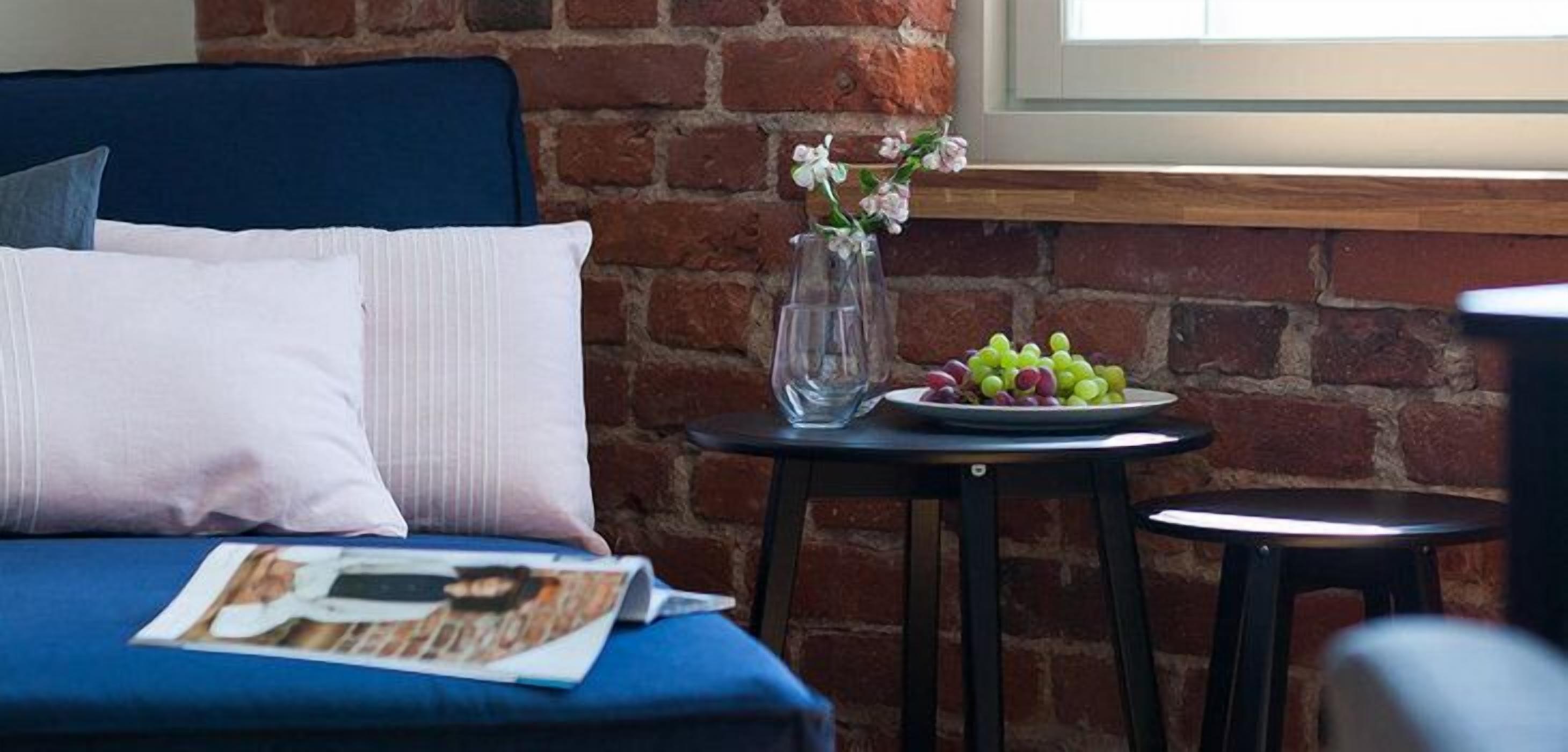 A bedroom with a brick wall, a window, a bed with blue bedding, a pillow, a magazine, a vase with flowers, and a plate of grapes on a small table.