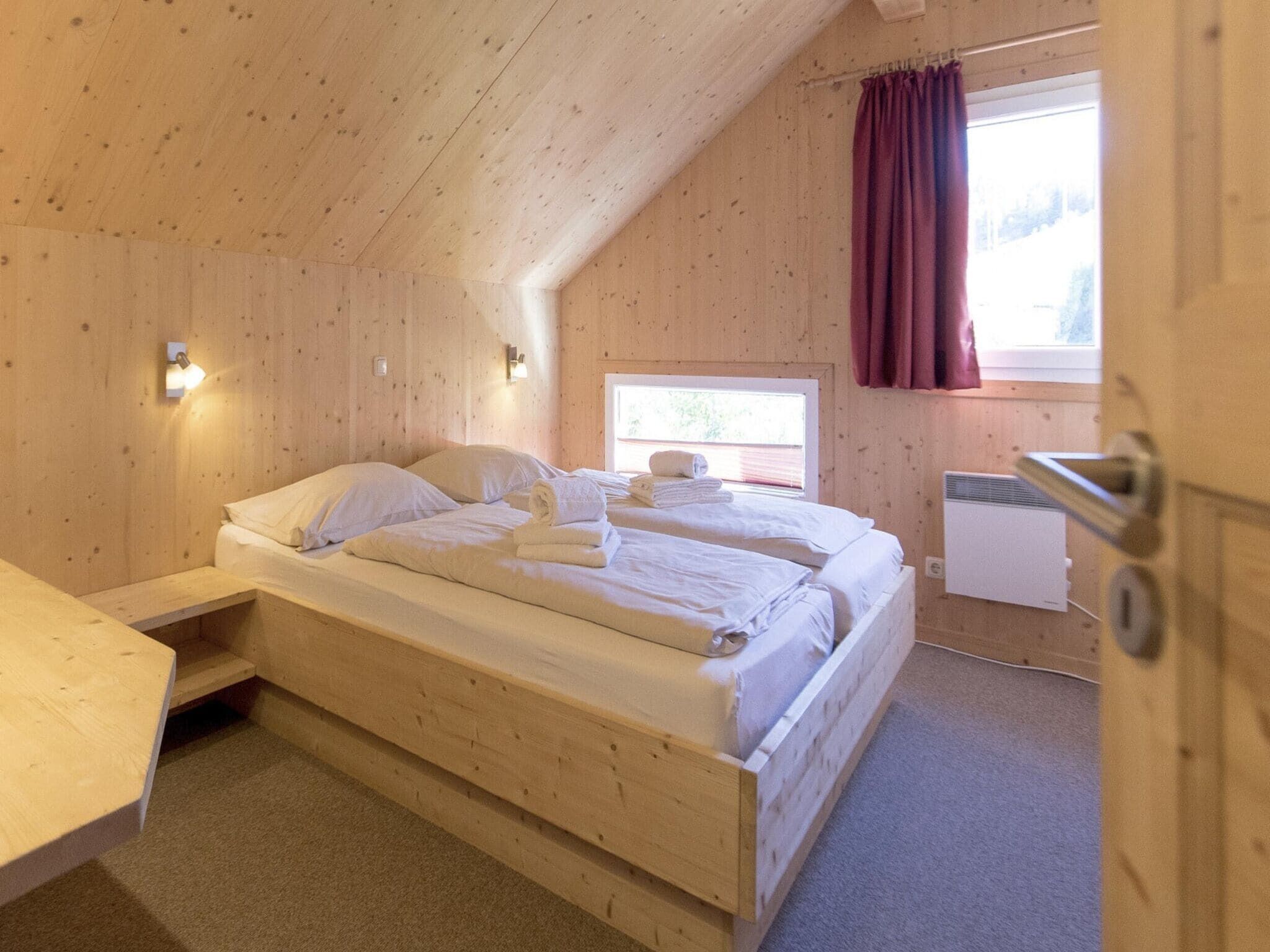 Un dormitorio de cabaña de madera con una cama individual, una mesita pequeña y una ventana con cortinas.