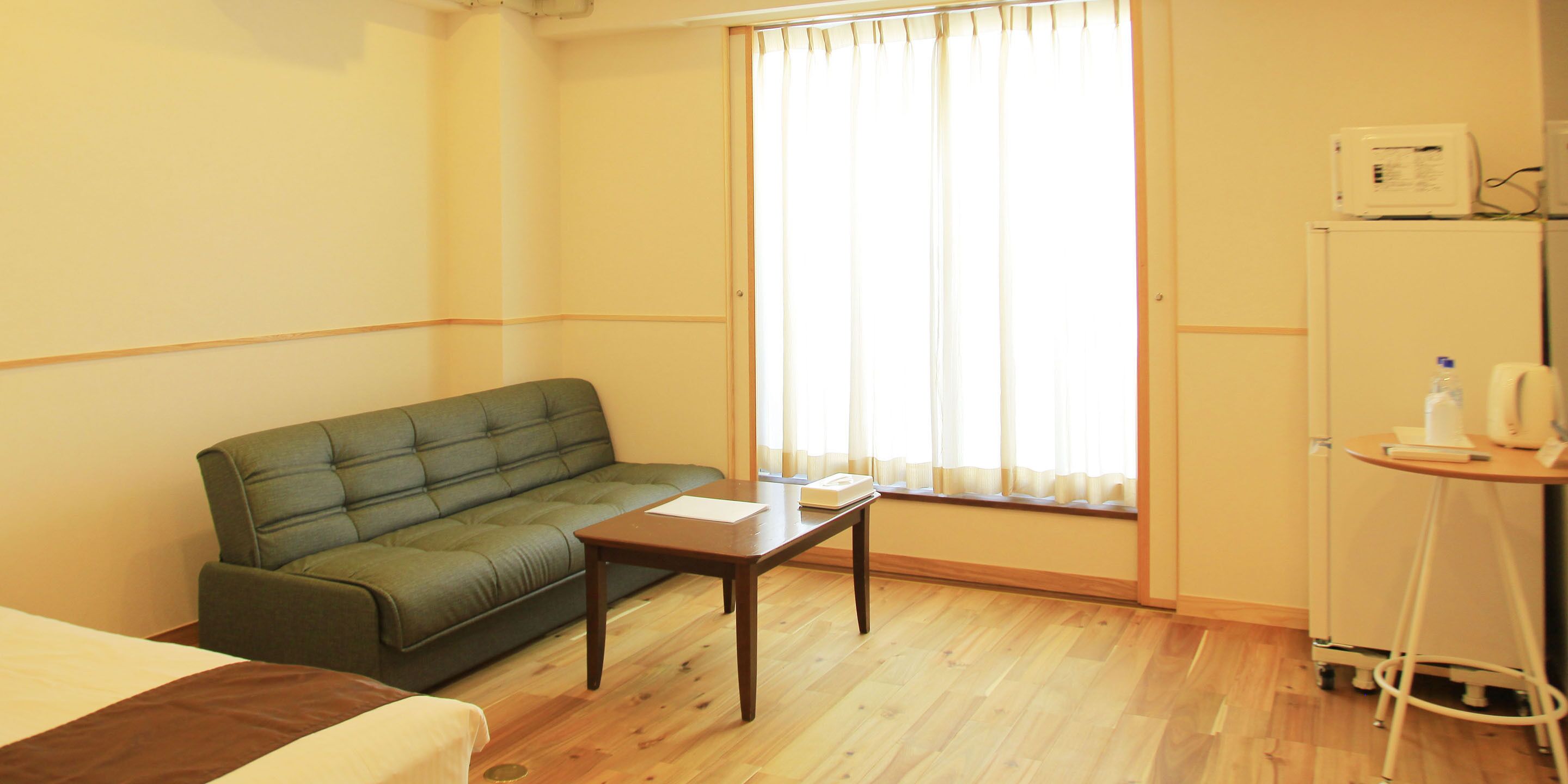 A hotel room with a green sofa, a wooden table, a bed, a window with curtains, and a minibar.