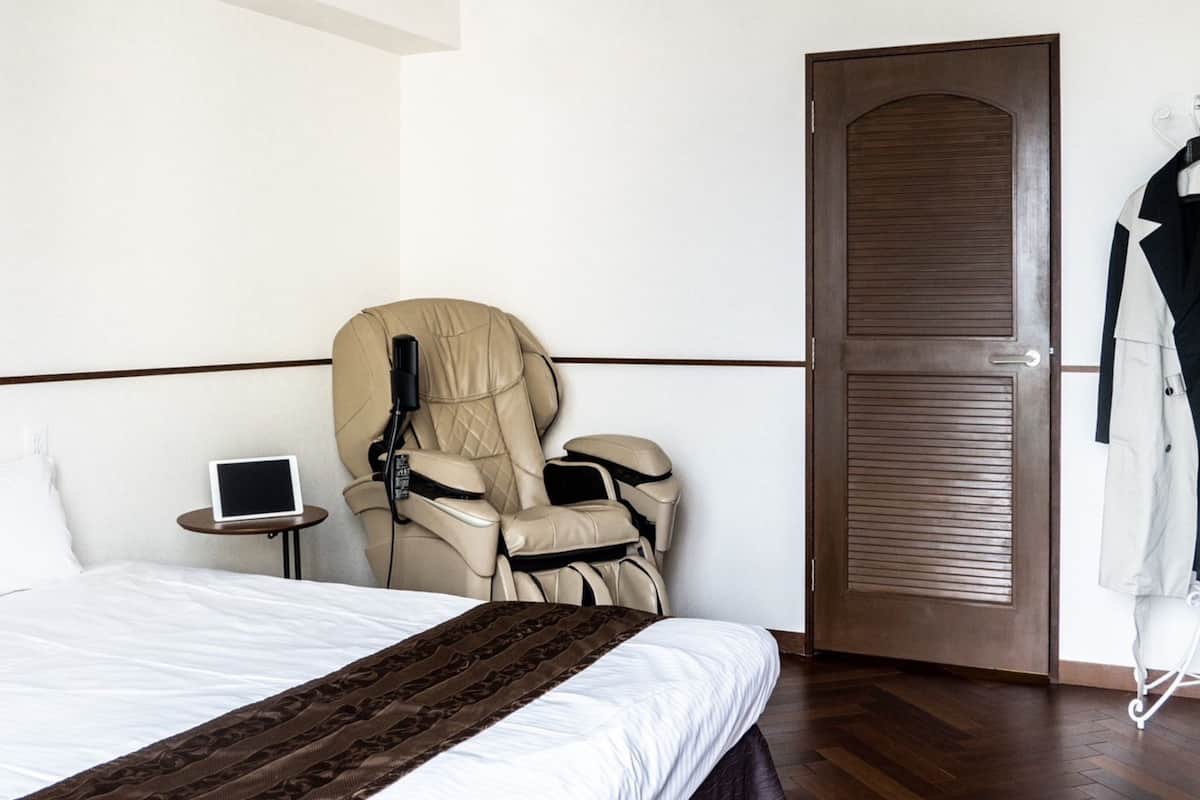 A hotel room with a bed, a massage chair, a small table with a tablet, and a coat rack.