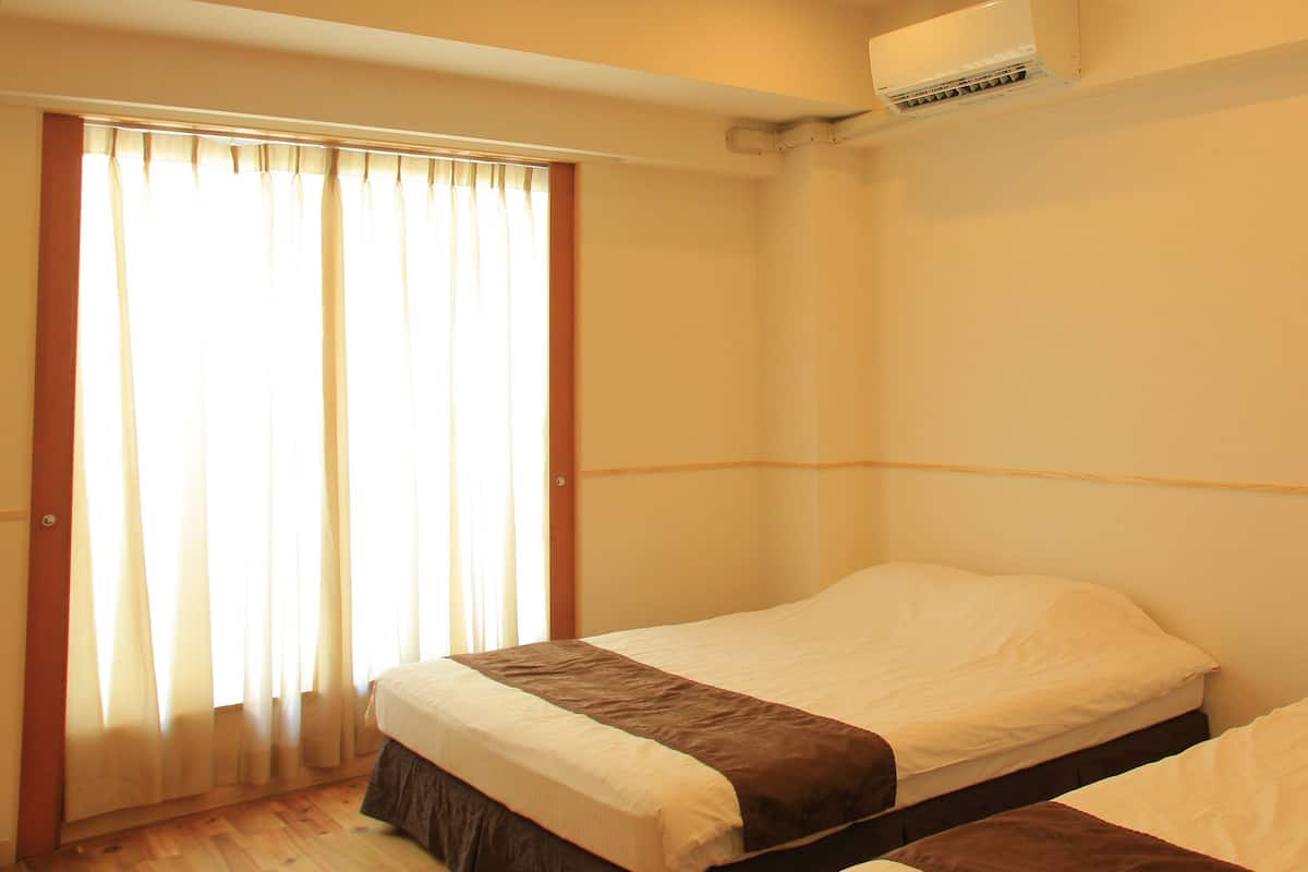 A bedroom with two single beds, a window with curtains, and a wall-mounted air conditioning unit.