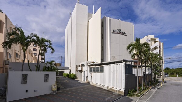 Un hotel moderno con un llamativo letrero que dice 'Sunset Terrace', un estacionamiento con acceso restringido y palmeras.
