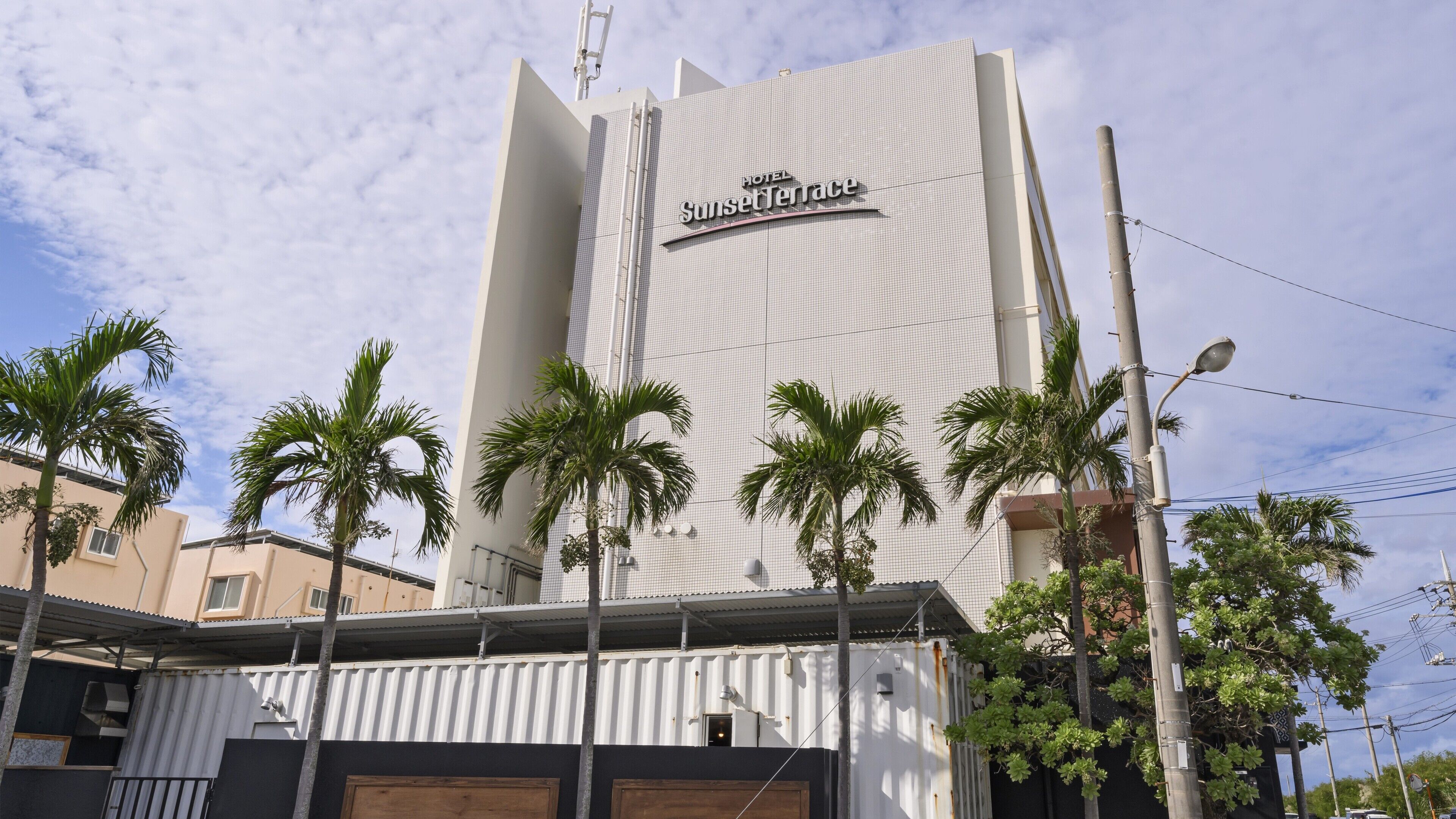 A hotel named Sunset Terrace with a modern design, featuring a prominent sign and a series of palm trees in front.