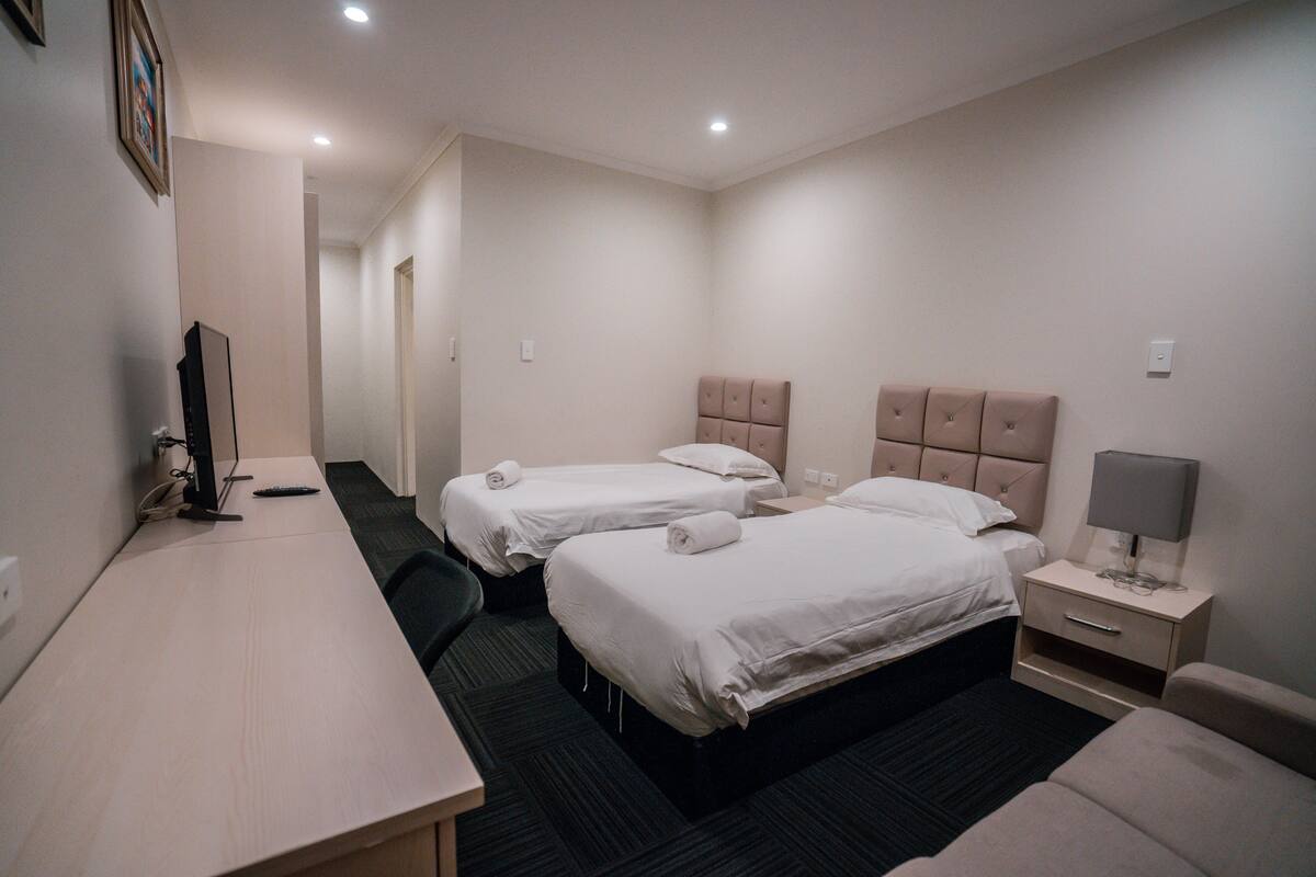A hotel room with two beds, a desk, a chair, a nightstand with a lamp, and a television.