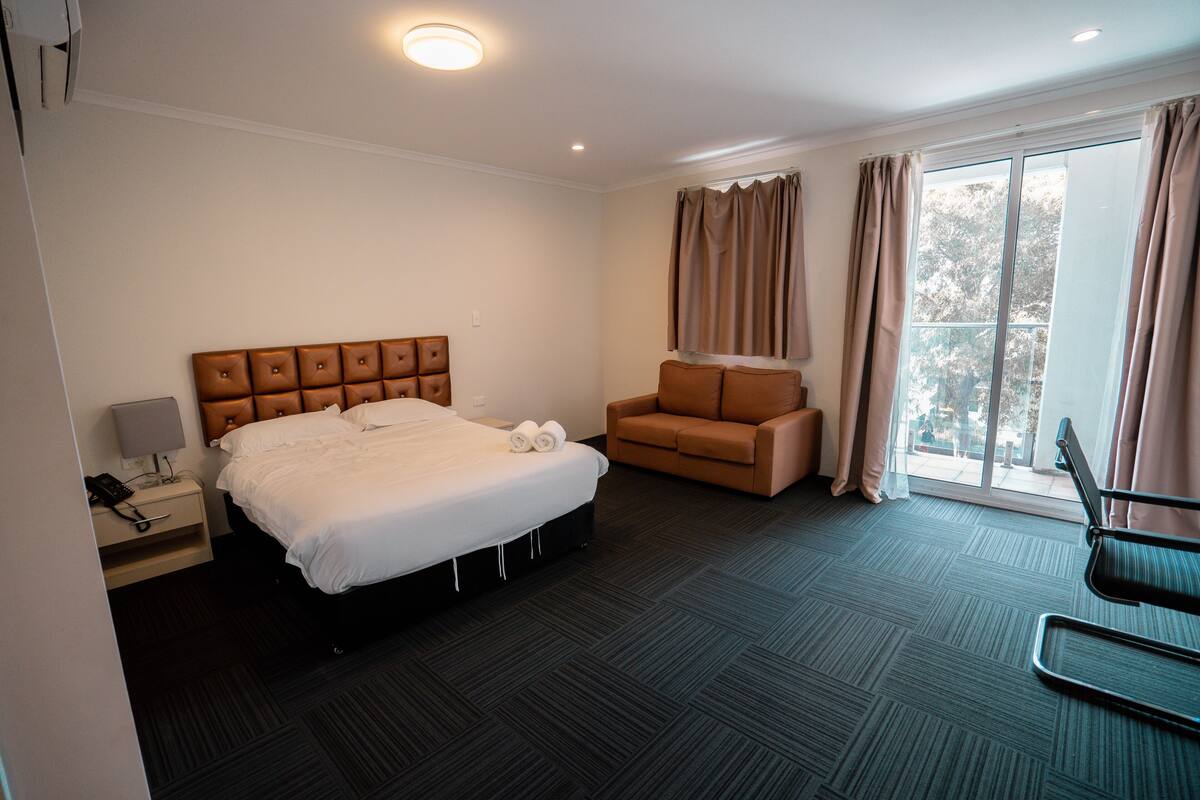 A hotel room with a large bed, a sofa, a desk with a lamp, and a view of the outdoors through a window.