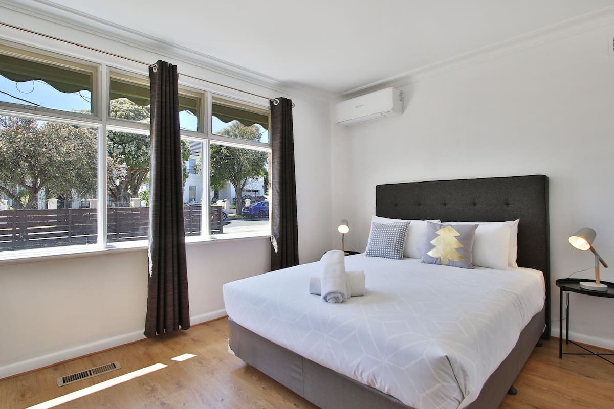 Una cama bien hecha con cabecera gris y ropa de cama blanca, una mesita de noche con lámpara y una ventana con cortinas.