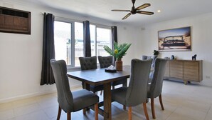 A dining area with a wooden table, grey upholstered chairs, a ceiling fan, and a window with curtains. - Ivy Units Glen Waverley (Glen Waverley)