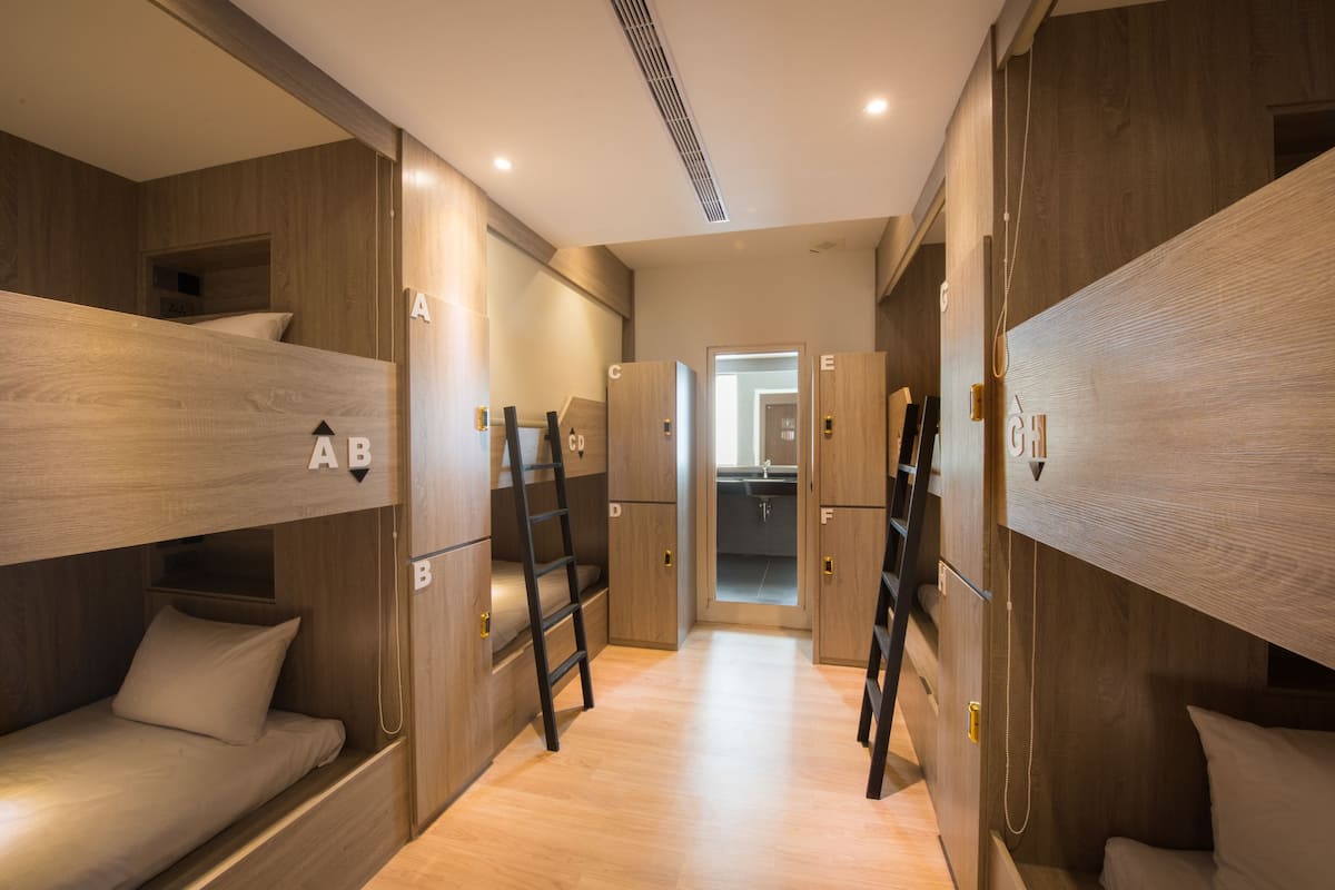 A compact, modern room with bunk beds labeled A to G, wooden walls, and a central hallway leading to a bathroom.