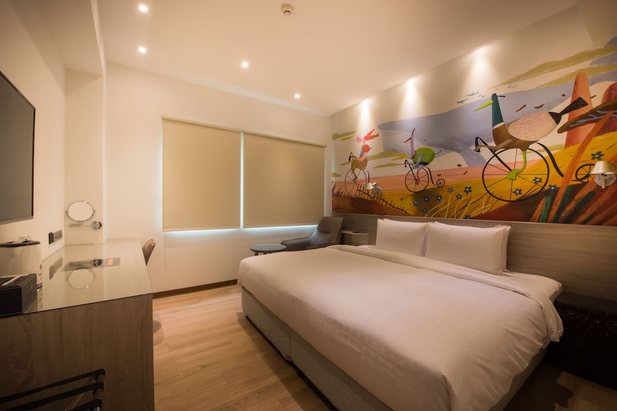 A modern hotel room with a large bed, a flat-screen TV, a small bedside table, and a window with blinds.