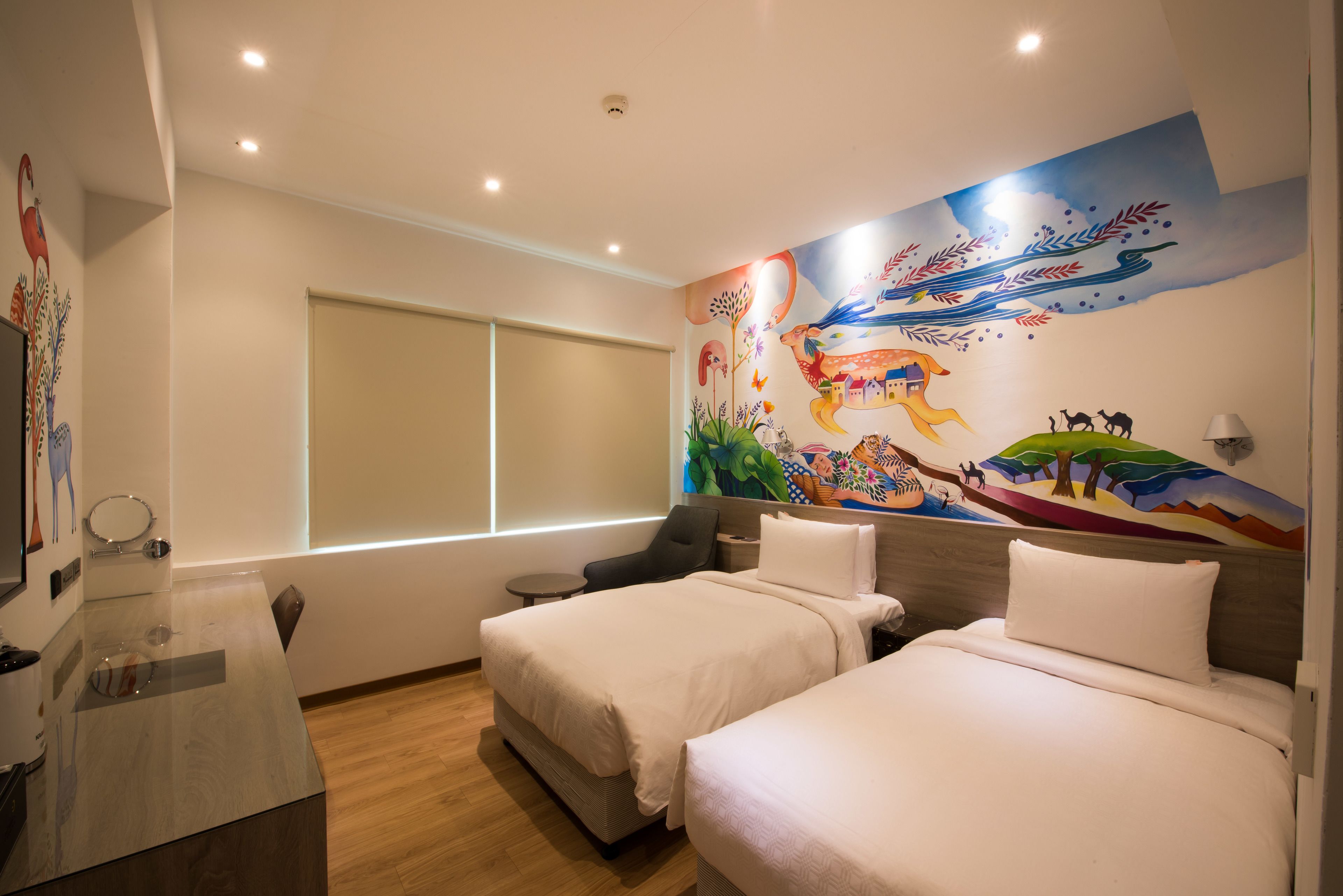 A hotel room with two beds, a large mural on the wall, a window, and a desk with a lamp.
