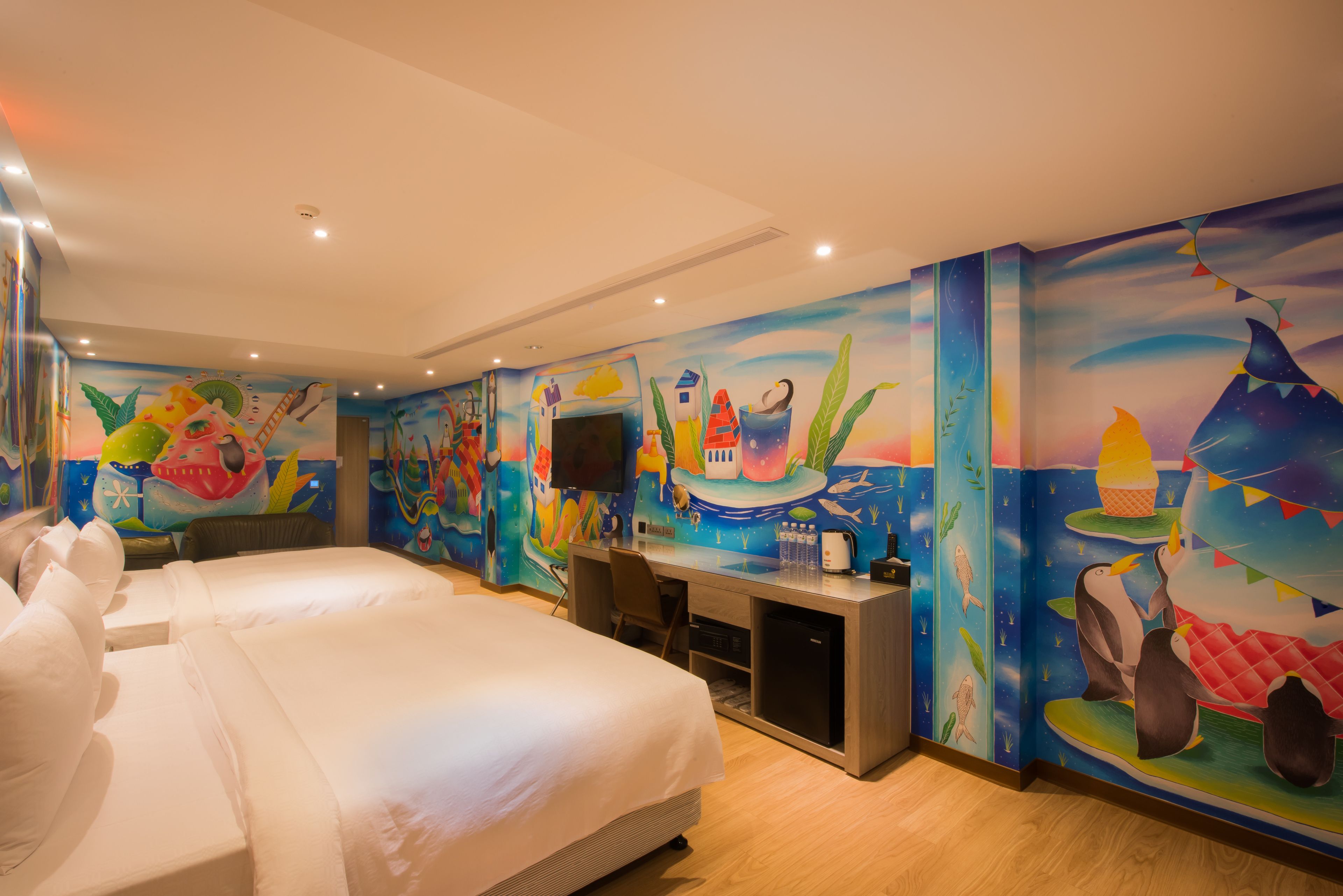 Ein Hotelzimmer mit zwei Betten, einem Schreibtisch, einem Fernseher und farbenfrohen Wandgemälden.