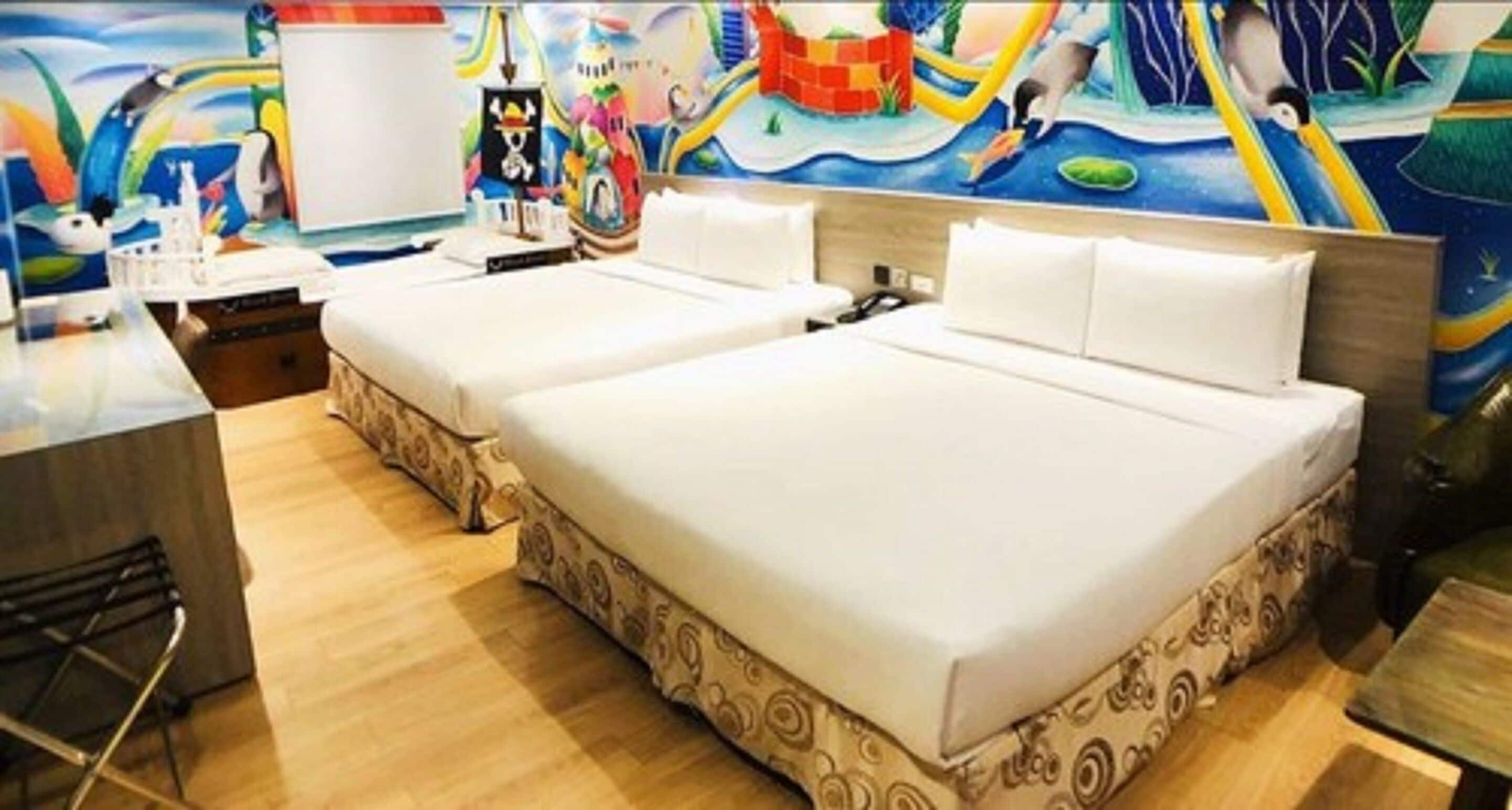 A hotel room with two beds, a wooden floor, a colorful mural on the wall, and a small table with a chair.