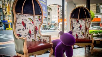 Interior de hotel con dos sillas ornamentadas con estampados de pájaros, un cojín con forma de oso morado y un suelo de baldosas.