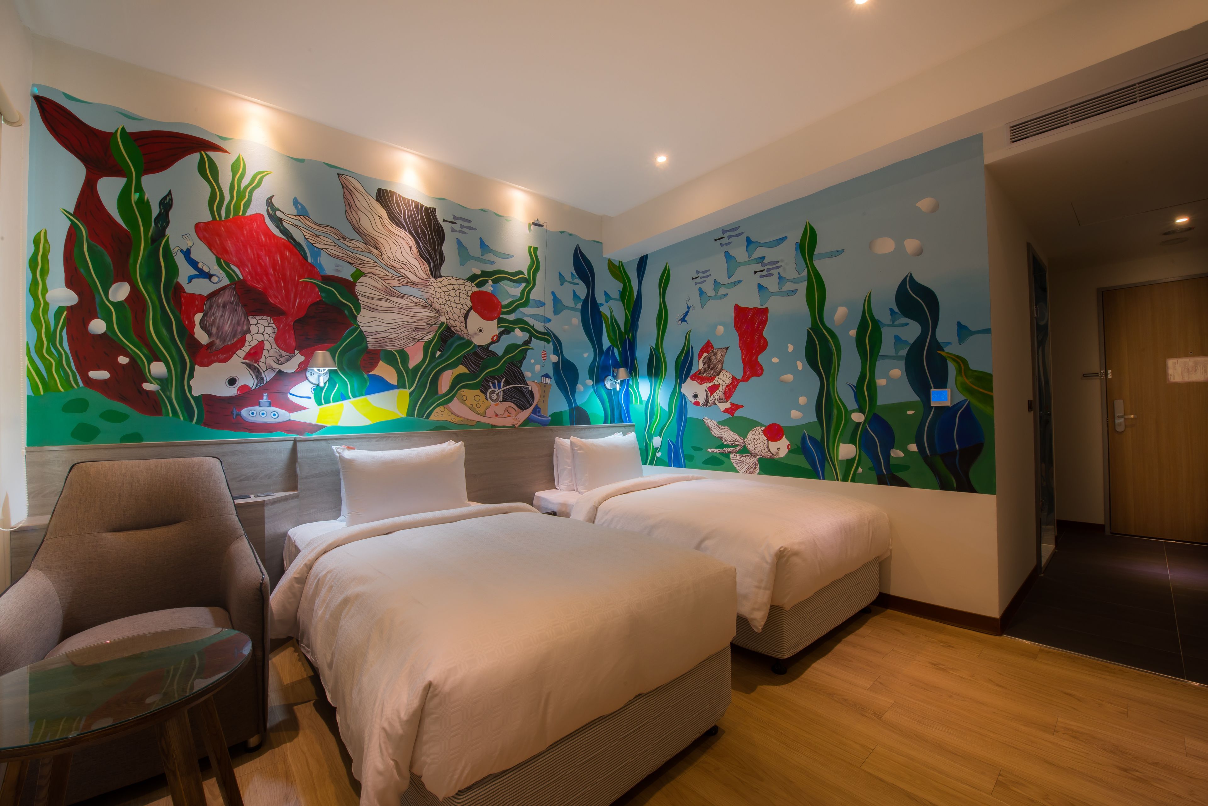 A hotel room with two beds, a chair, a small table, and a mural of fish and seaweed on the wall.