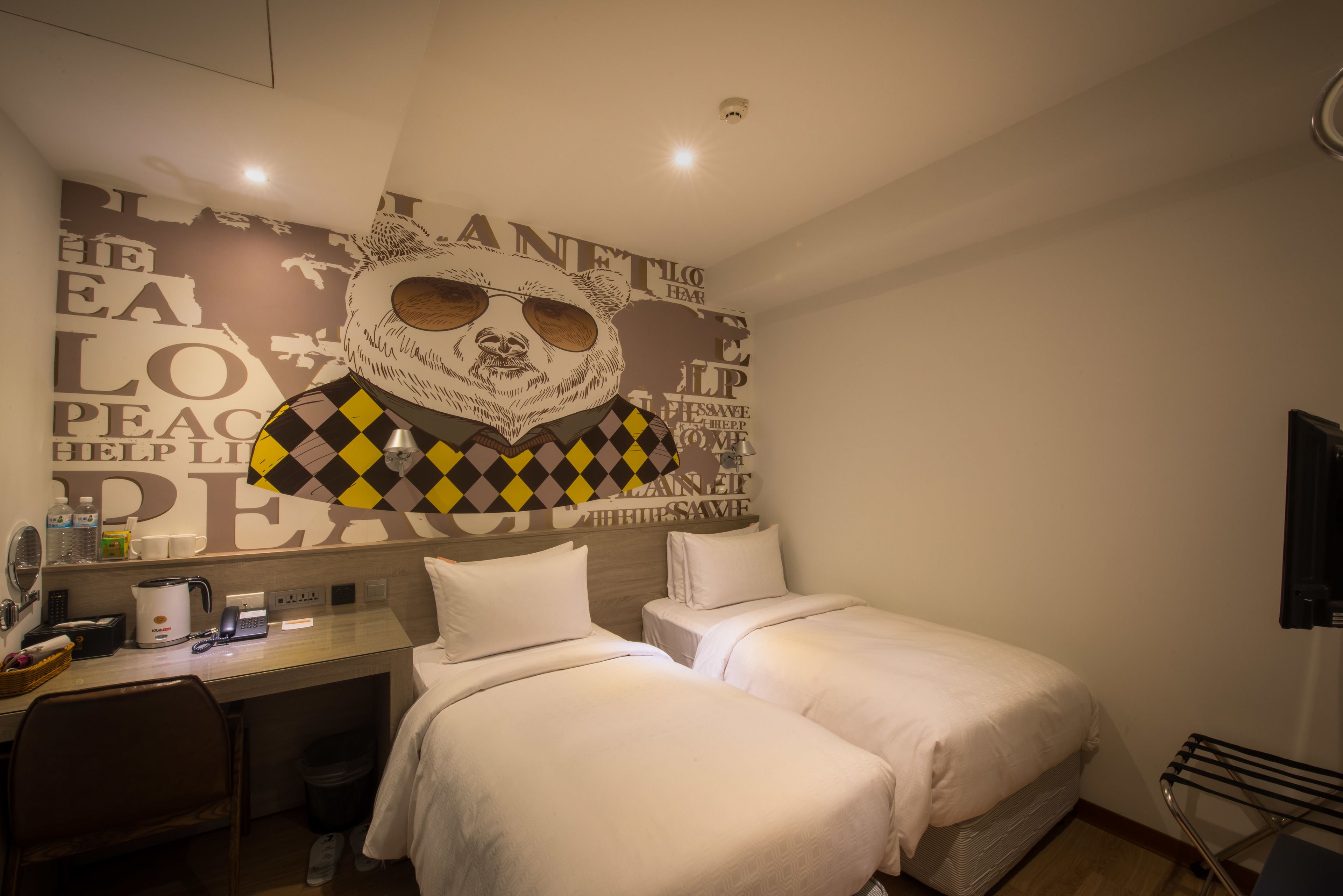 A hotel room with two beds, a desk with a kettle, a mirror, and a wall with a large bear illustration.