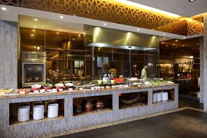 A buffet setup with various food items and utensils, a large kitchen area in the background, and a patterned wall design. - Shixishu Jianguo Yalong Bay Resort Hotel (Sanya)