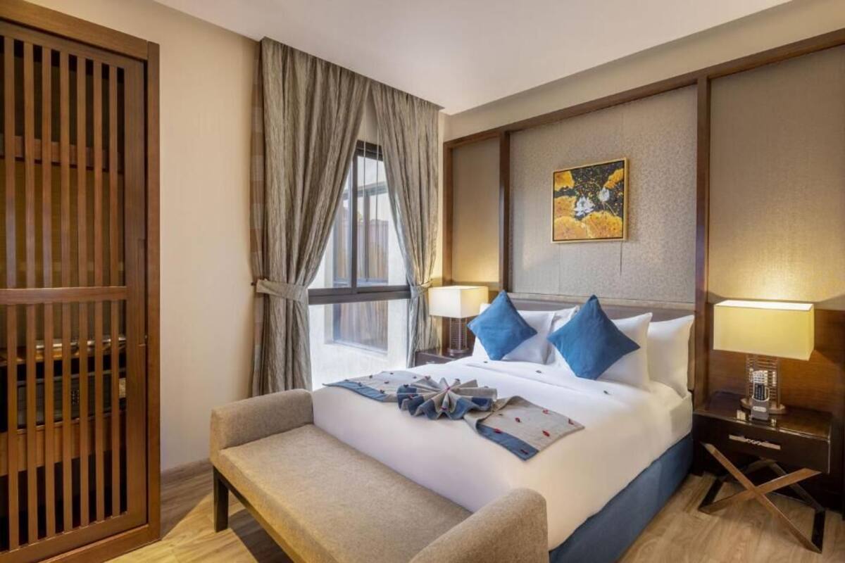 A hotel room with a bed, a bedside table with a lamp, a chair, a window with curtains, and a wooden partition.