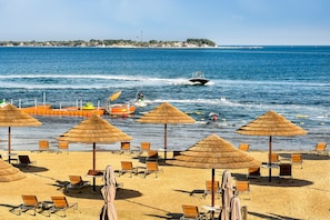 A beach with rows of sun loungers and thatched umbrellas, a jet ski in the water, and a distant shoreline with buildings. - Braira Al azizia Hotel (Al Khobar)