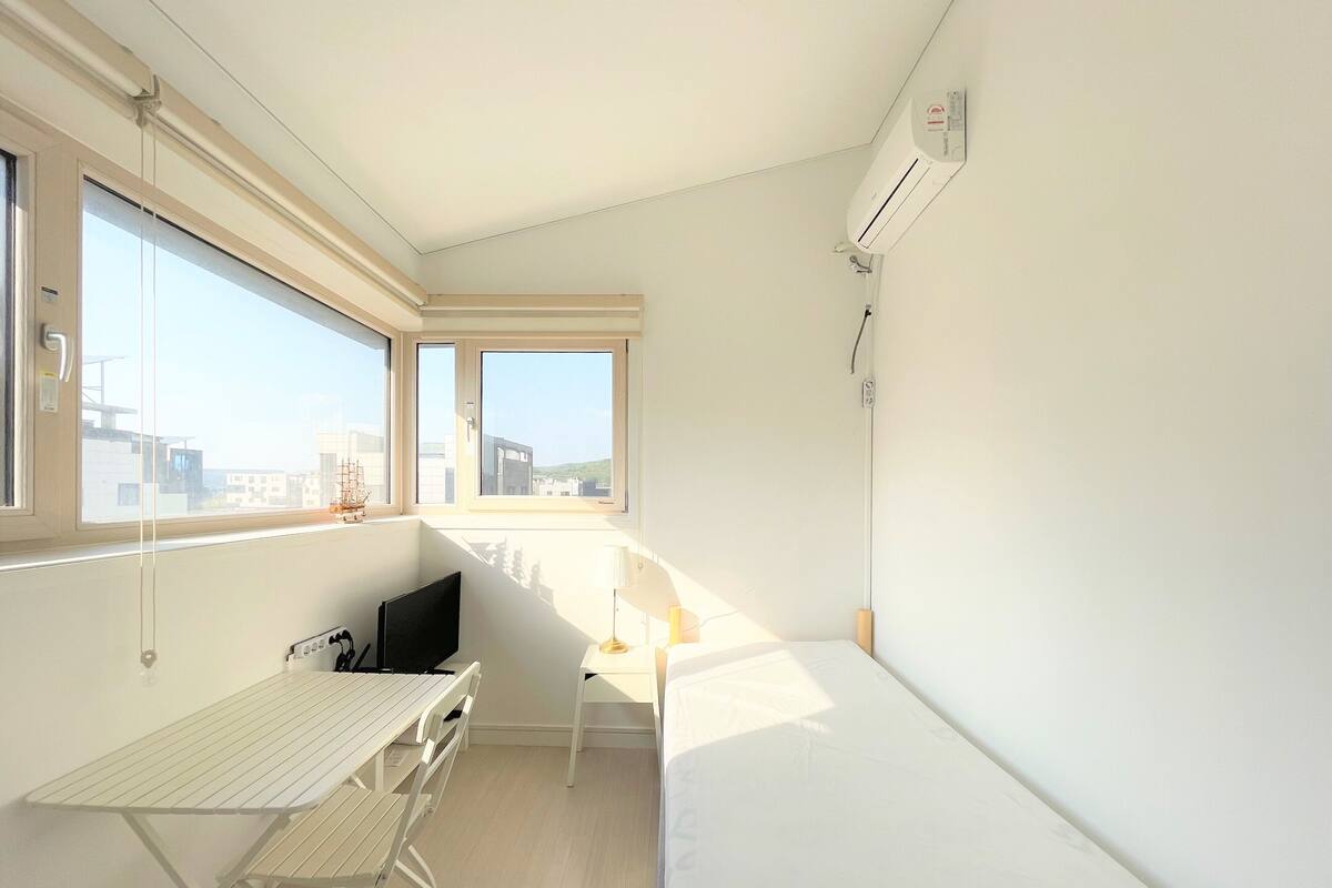 Quarto pequeno e luminoso com uma cama, uma mesa e uma cadeira. Janelas amplas oferecem vista para edifícios lá fora.
