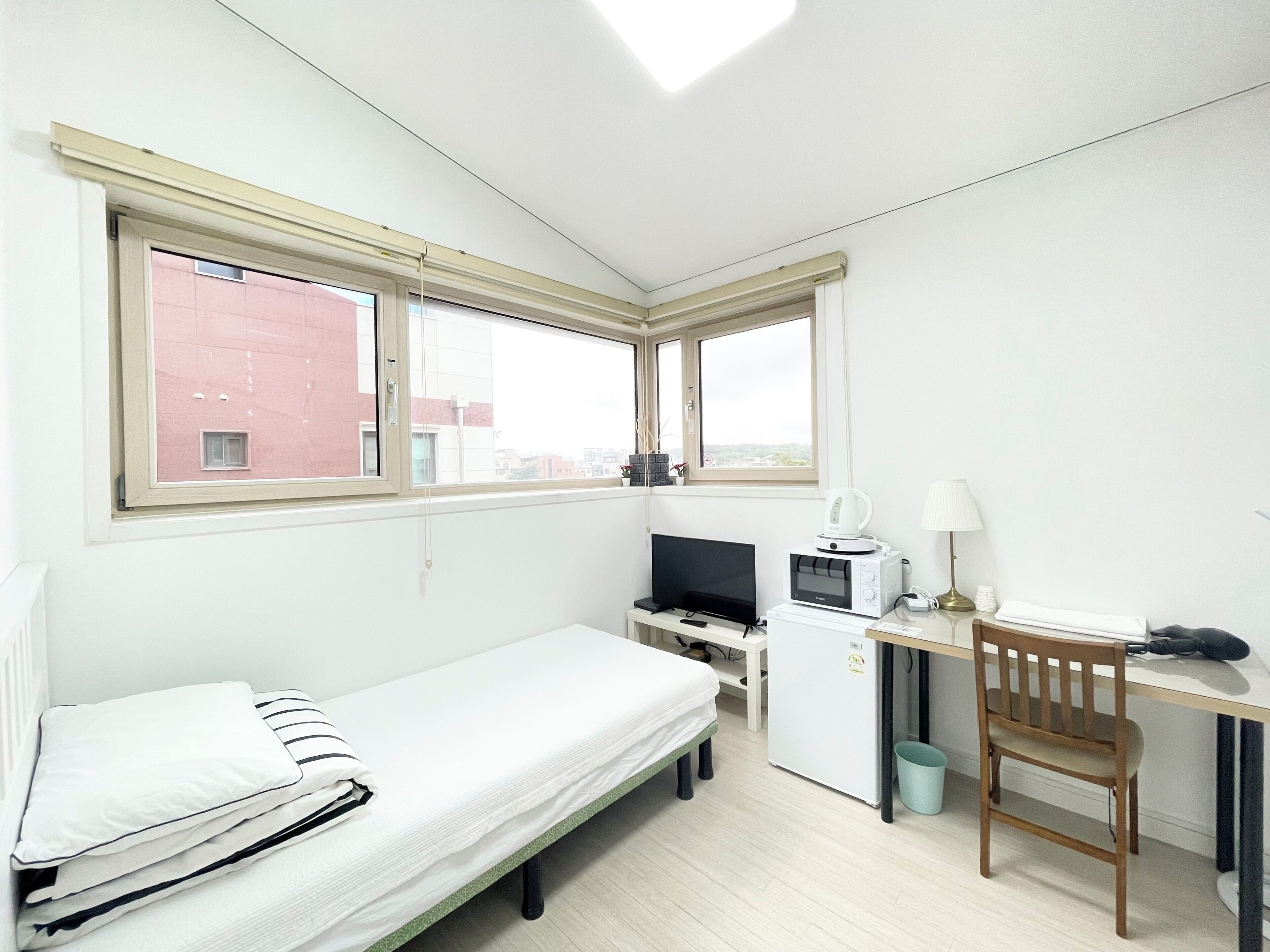 Une petite pièce bien éclairée, avec un lit simple, un bureau équipé d’un ordinateur et d’un micro-ondes, une chaise et une fenêtre donnant sur des immeubles.