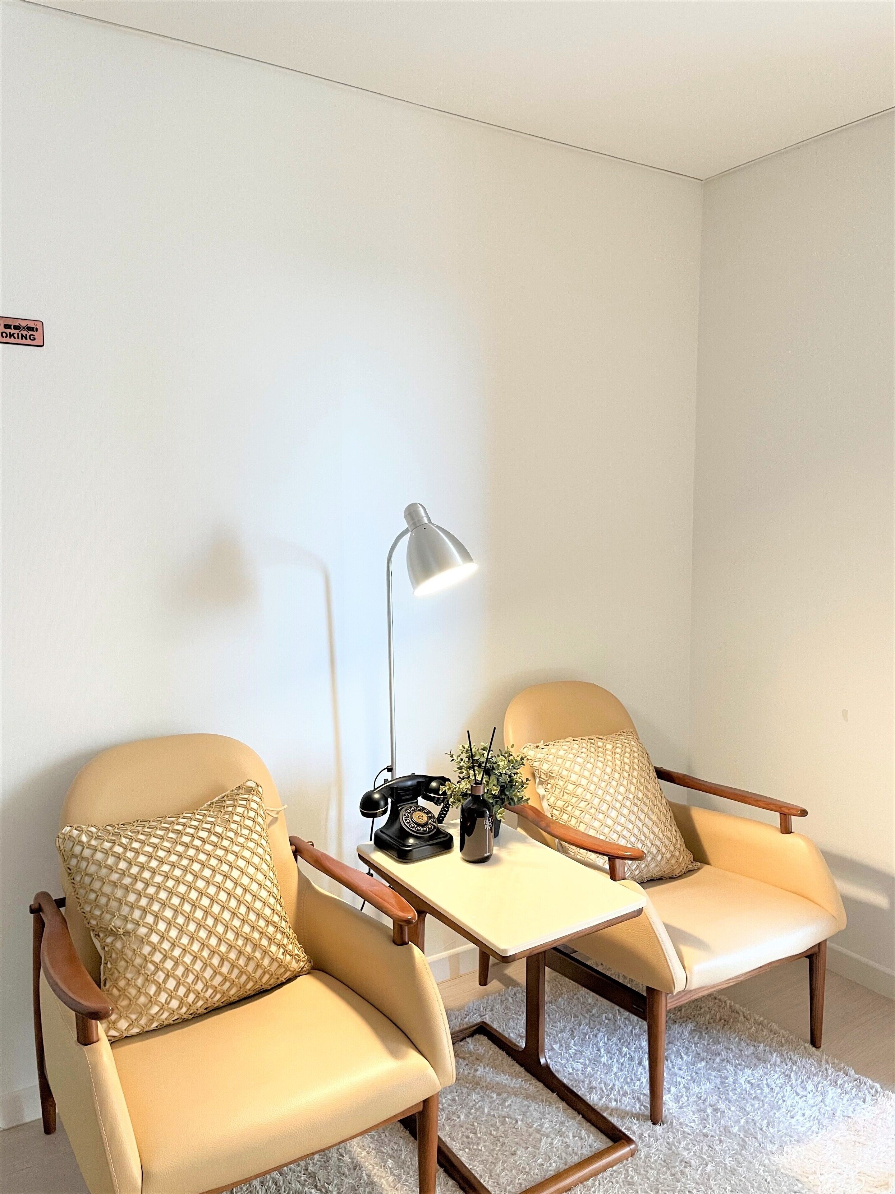 A room with two beige armchairs, a small table with a lamp, a telephone, and a plant.